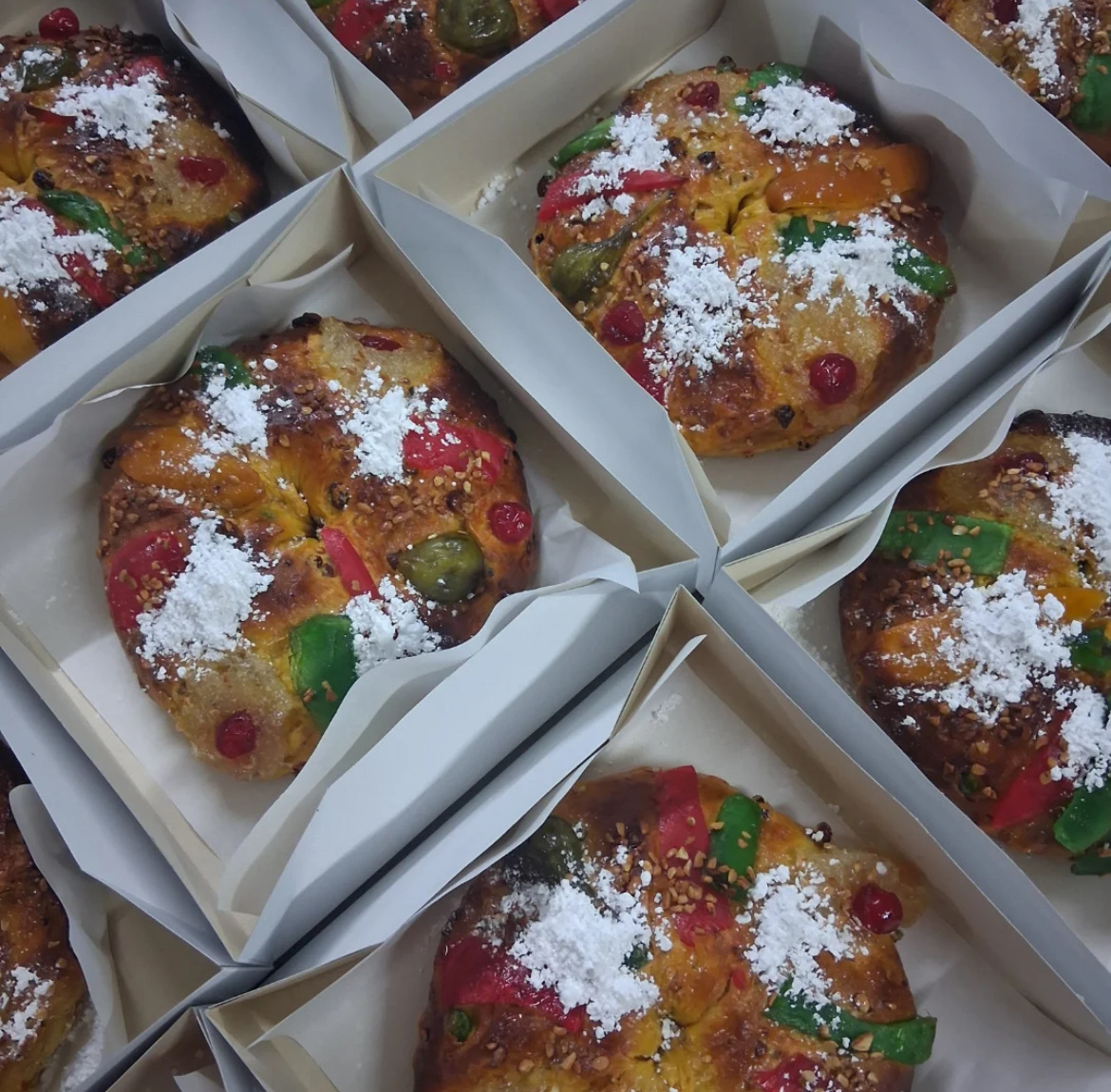 A close-up shot of several Bolo Rei cakes, a traditional Portuguese Christmas treat. The cakes are round, topped with candied fruits and powdered sugar, and presented in individual boxes.