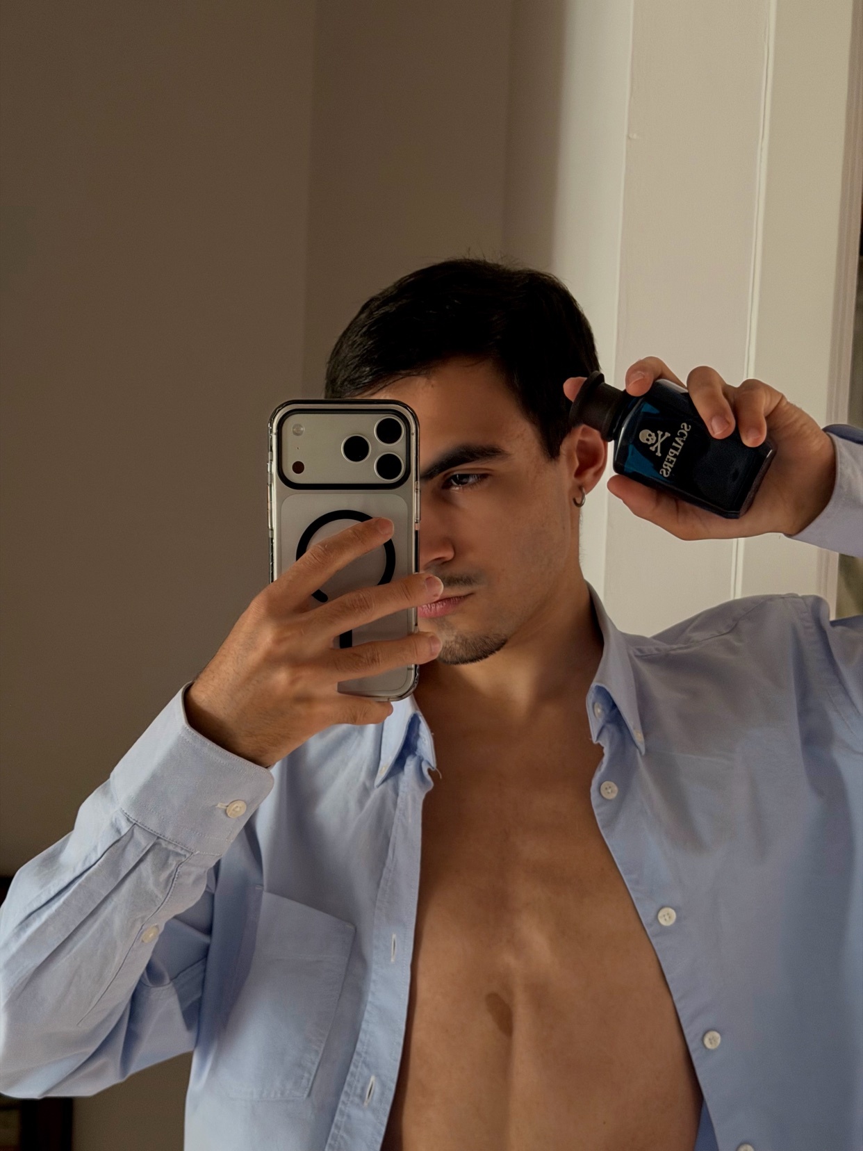 A man holds a bottle of cologne while taking a mirror selfie. He is wearing an unbuttoned blue dress shirt, revealing his bare chest.