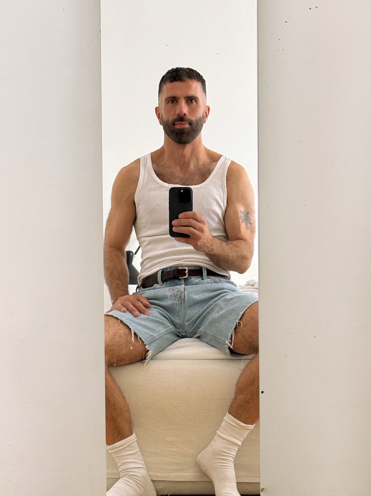 A man with a beard and a tattoo on his arm takes a mirror selfie while wearing a white tank top, denim shorts, and white socks. He is sitting down and holding his phone to take the picture.