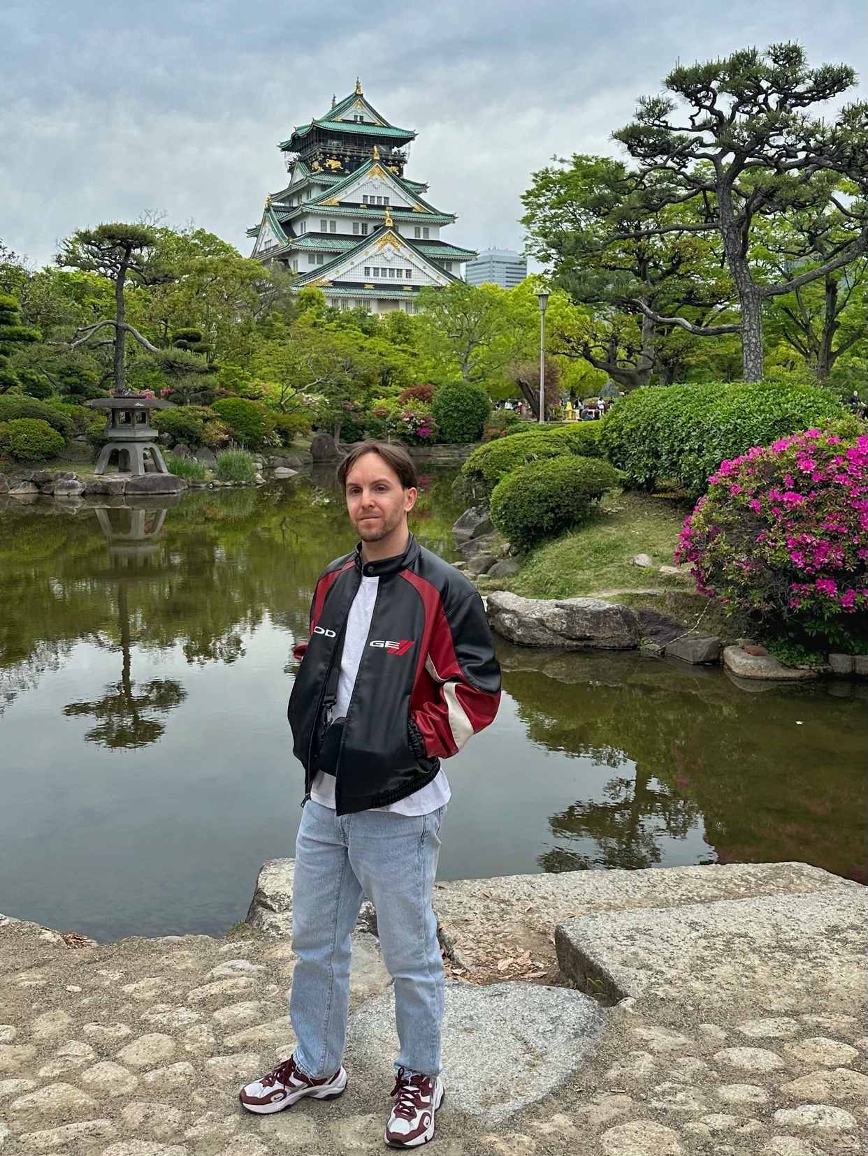 A man stands in front of a Japanese castle with a pond and manicured gardens in the foreground. The castle has a distinctive green roof and white walls, set against a cloudy sky.