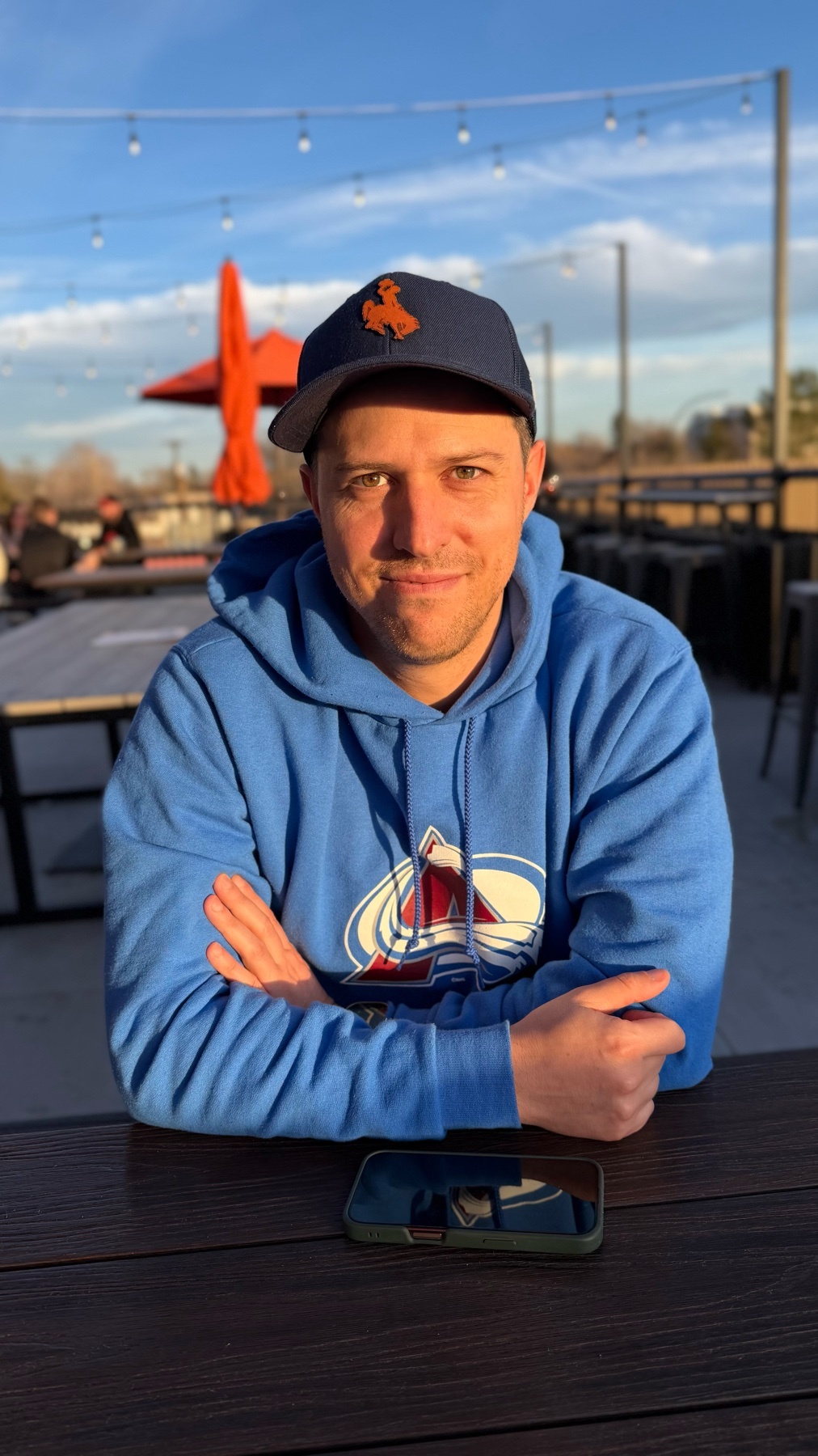 A man sits at a table outdoors, smiling at the camera. He wears a blue hoodie and a baseball cap.