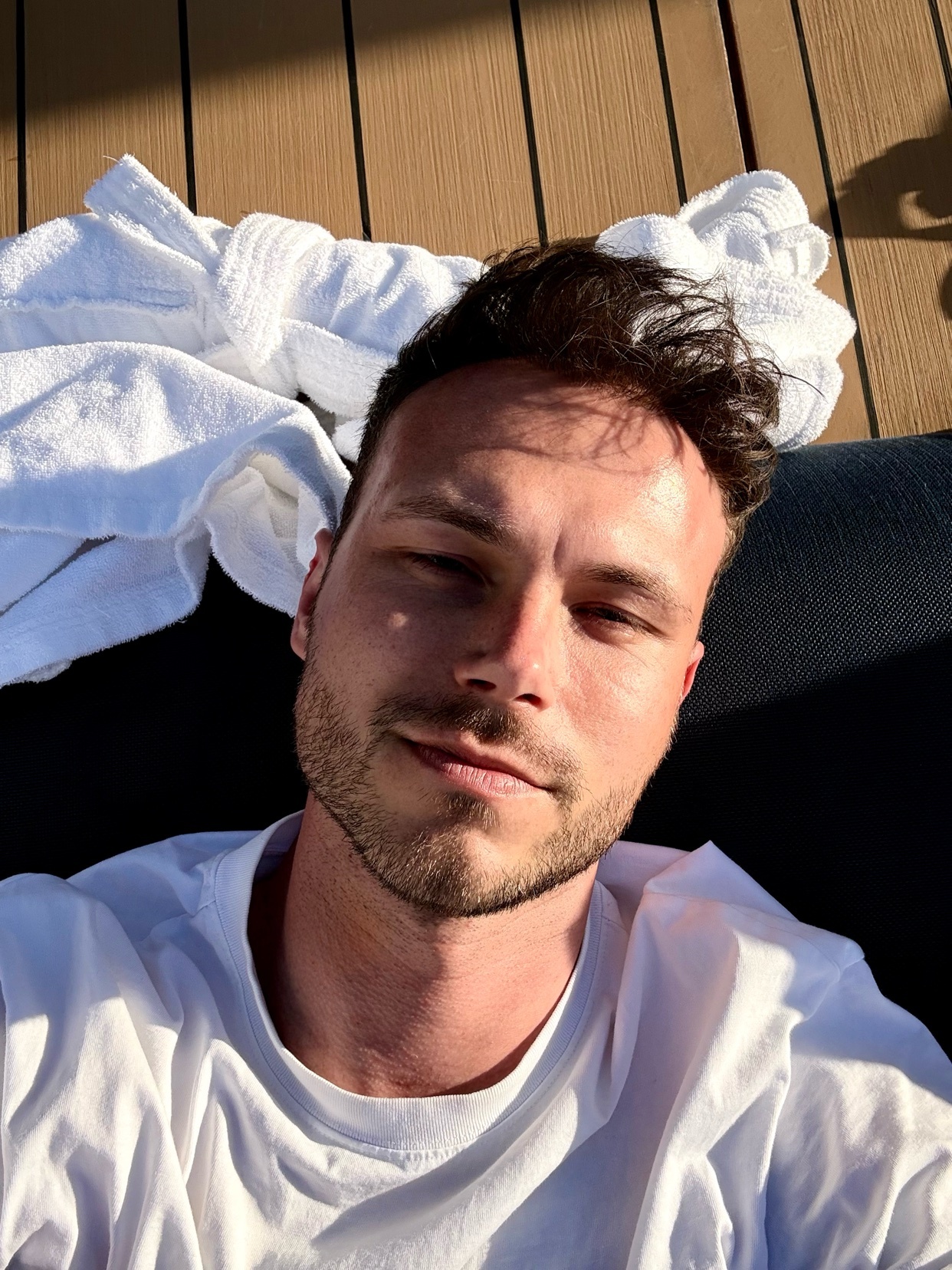 A man with stubble and dark hair takes a selfie while relaxing outdoors. The sun casts shadows on his face and a white robe is visible in the background.
