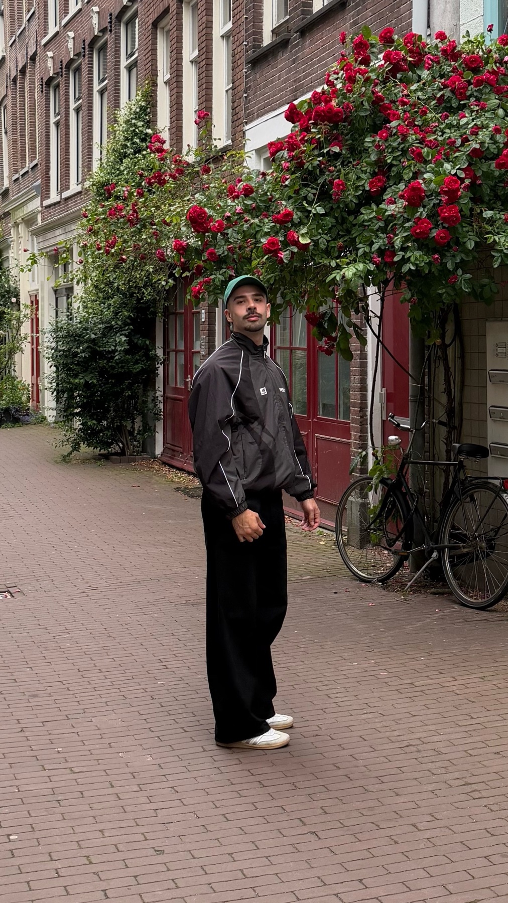 A man in a black tracksuit and a green baseball cap stands on a cobblestone street lined with red roses and historic buildings. A bicycle is parked nearby.