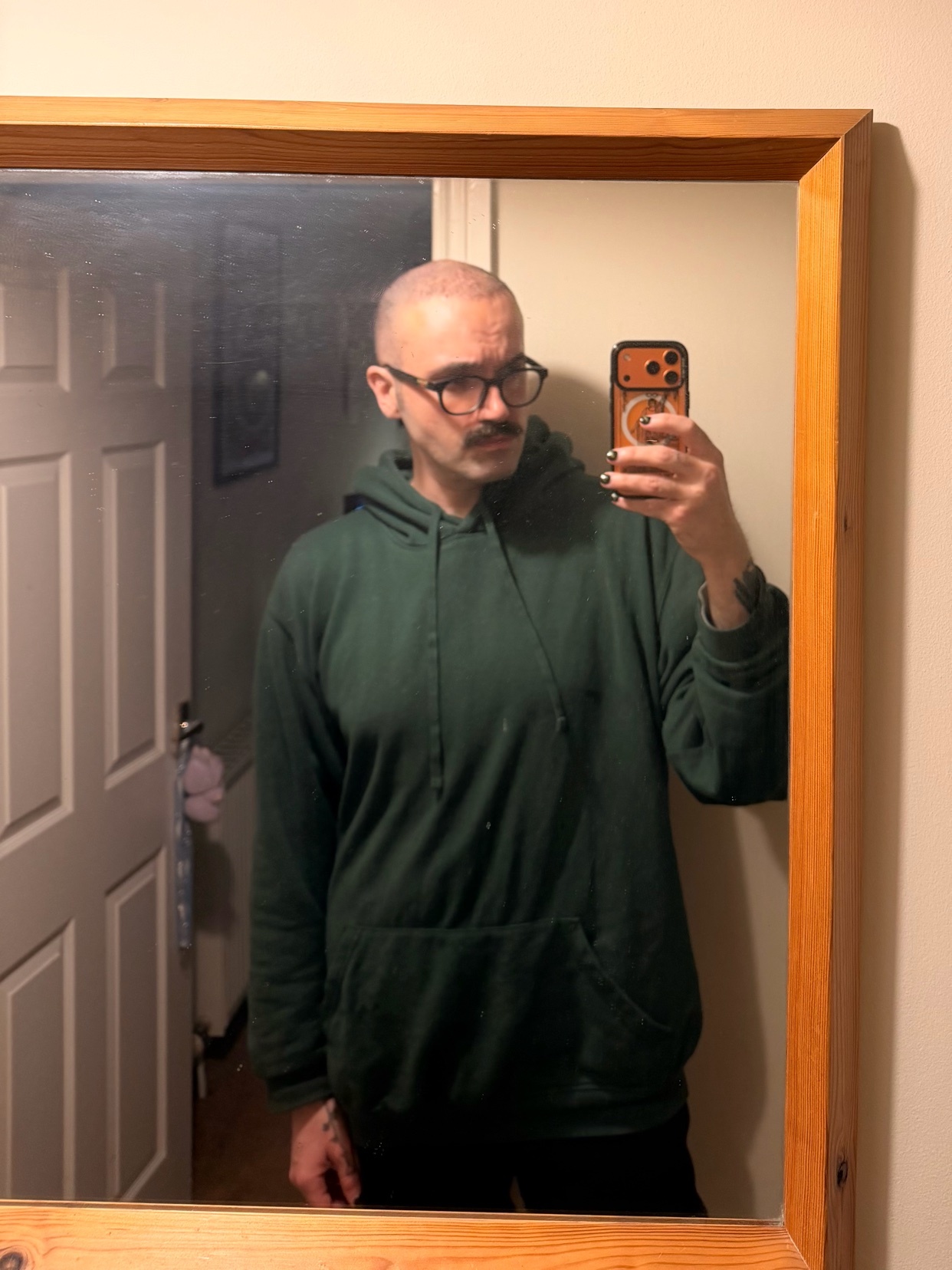A man with a bald head, glasses, and a mustache takes a mirror selfie in a dark green hoodie.