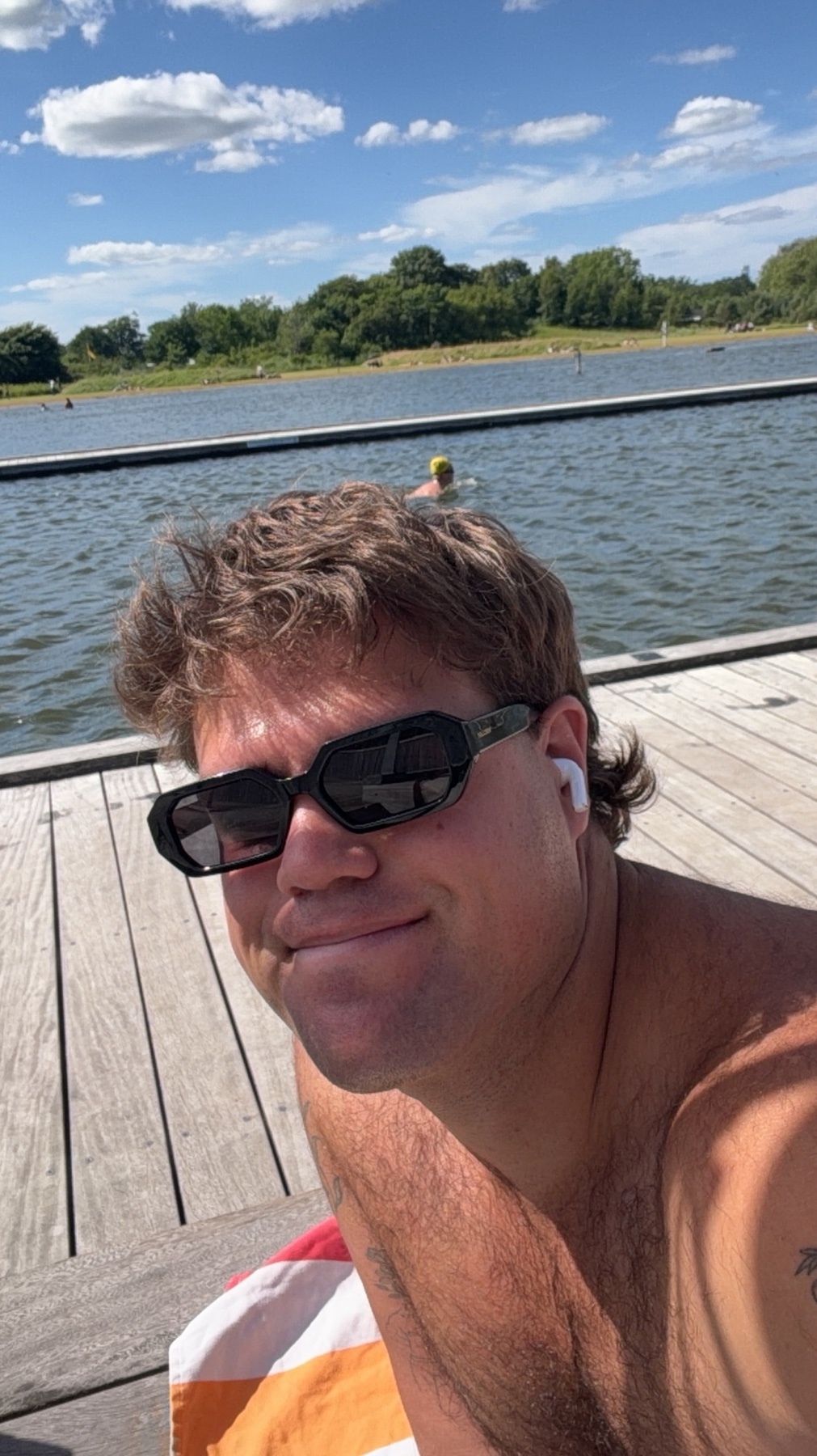 A man with a mullet and sunglasses smiles at the camera while sitting on a wooden pier next to a body of water. He is wearing earbuds and has a towel draped over his shoulder.