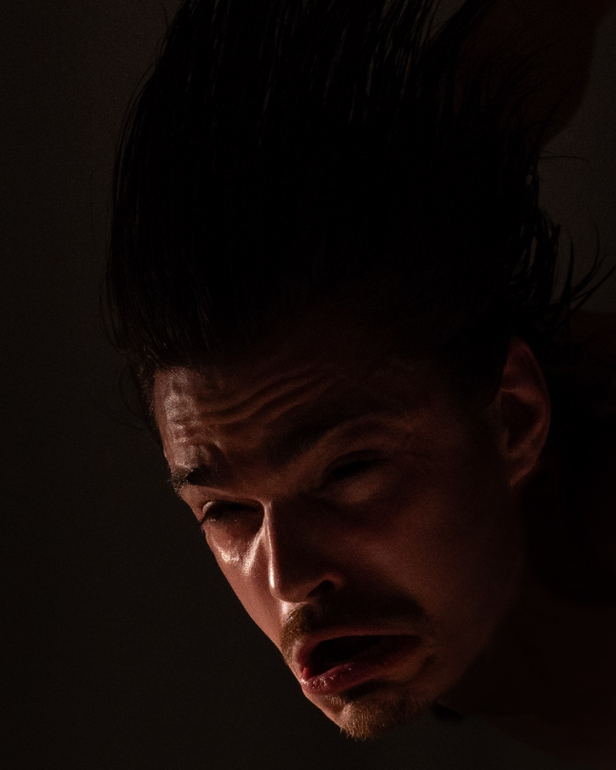 A man with dark, wet hair is captured in a dramatic, low-key portrait. His face is contorted in an intense expression, with his mouth slightly open and eyes wide.
