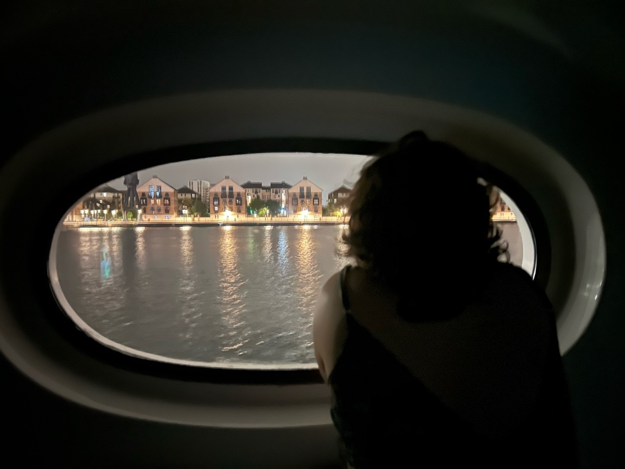 A person looks out of an oval window at a city skyline at night. The water reflects the lights of the buildings across the bay.