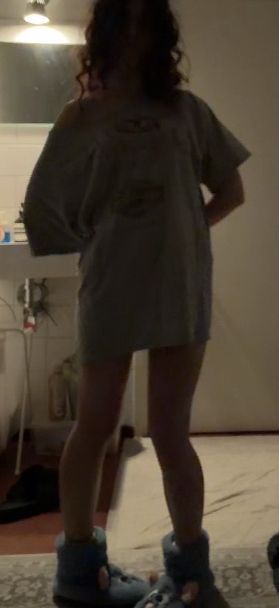 A woman with curly hair stands in an oversized t-shirt and fuzzy slippers. The image is dimly lit, with the woman's legs and torso visible.