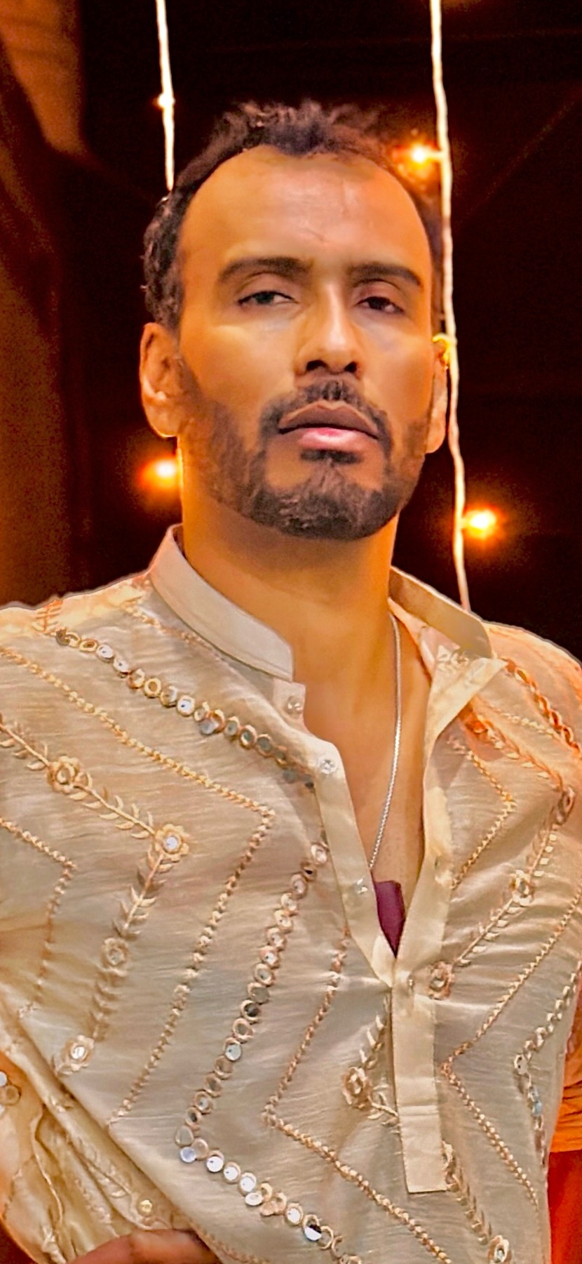 A man with a beard and stubble poses in a light-colored, embroidered ethnic outfit. He wears a silver chain around his neck, and the background is softly lit with string lights.