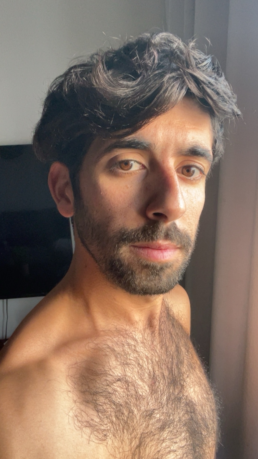 A close-up, chest-up shot of a man with dark, wavy hair and a beard. He has a hairy chest and is looking directly at the camera.