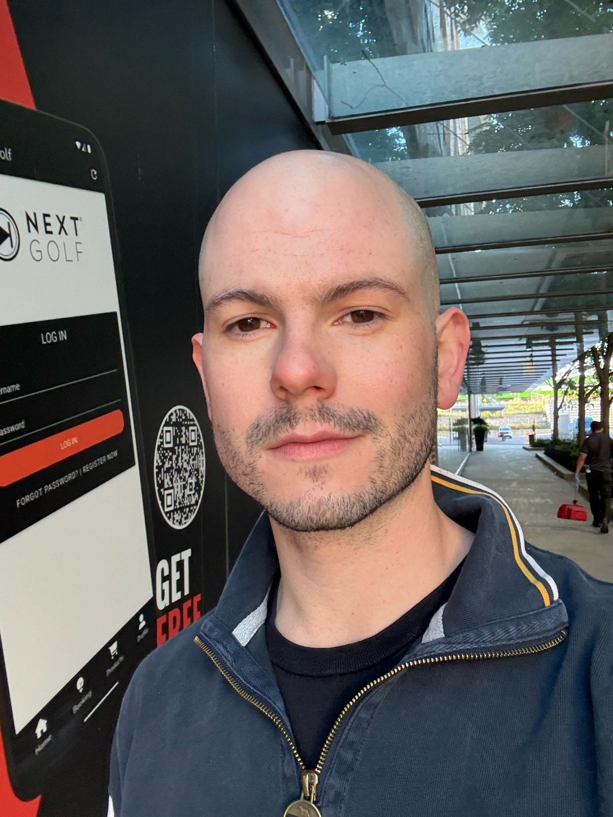 A man with a shaved head and stubble takes a selfie in front of a wall with a large advertisement for a golf app.