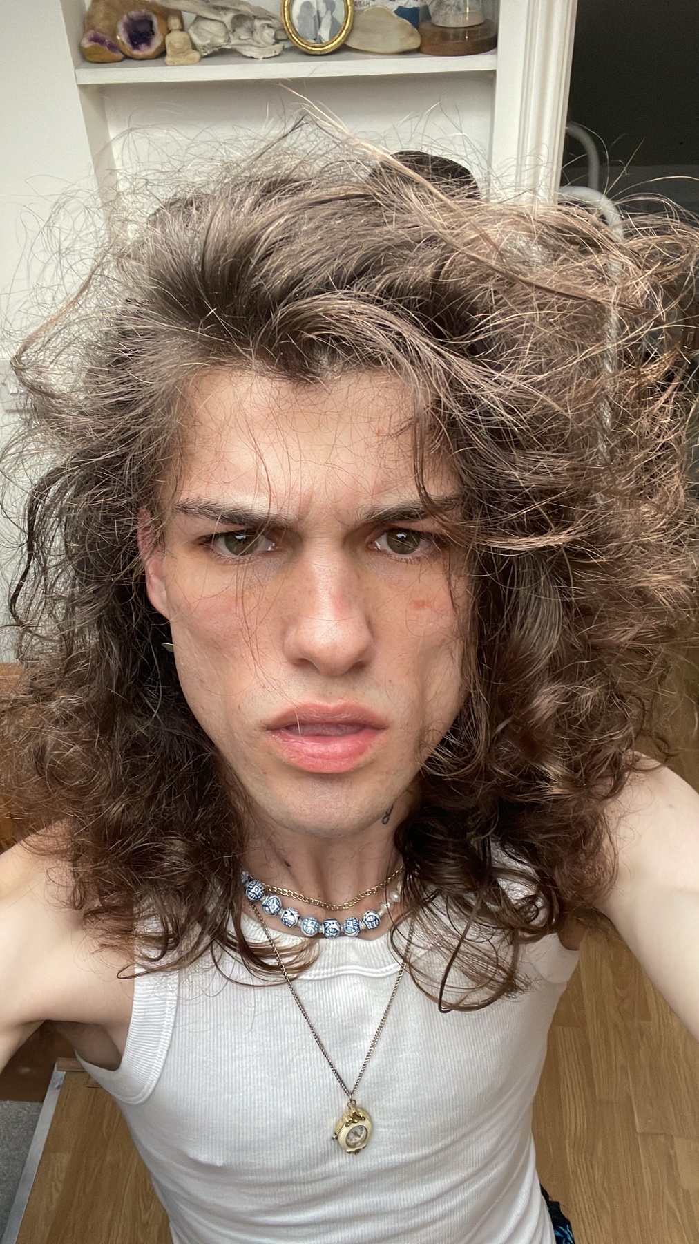 A selfie of a man with long, curly hair and a serious expression. He is shirtless, wearing a tank top and a necklace.