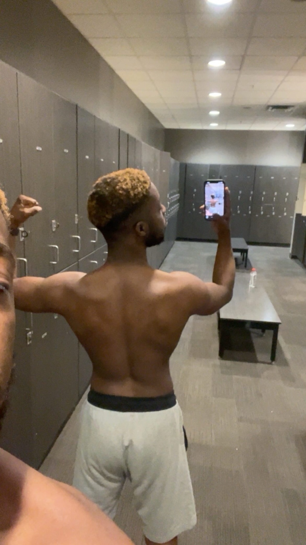 A man with blonde curly hair takes a selfie in a gym locker room, flexing his back muscles. He is wearing light-colored athletic shorts.