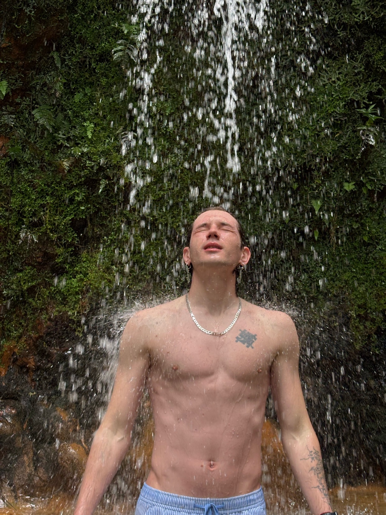 A shirtless man stands under a waterfall with his eyes closed and face tilted upwards. Lush green moss and rocks form the backdrop behind the cascading water.