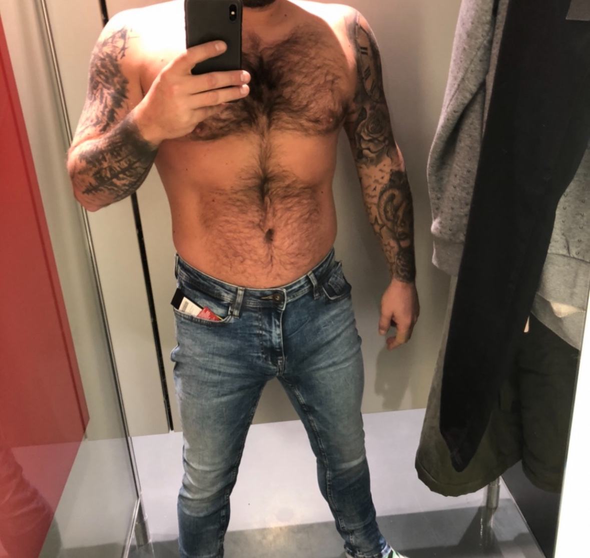 A man in a fitting room takes a mirror selfie showing off his tattooed arms and hairy chest while wearing jeans.