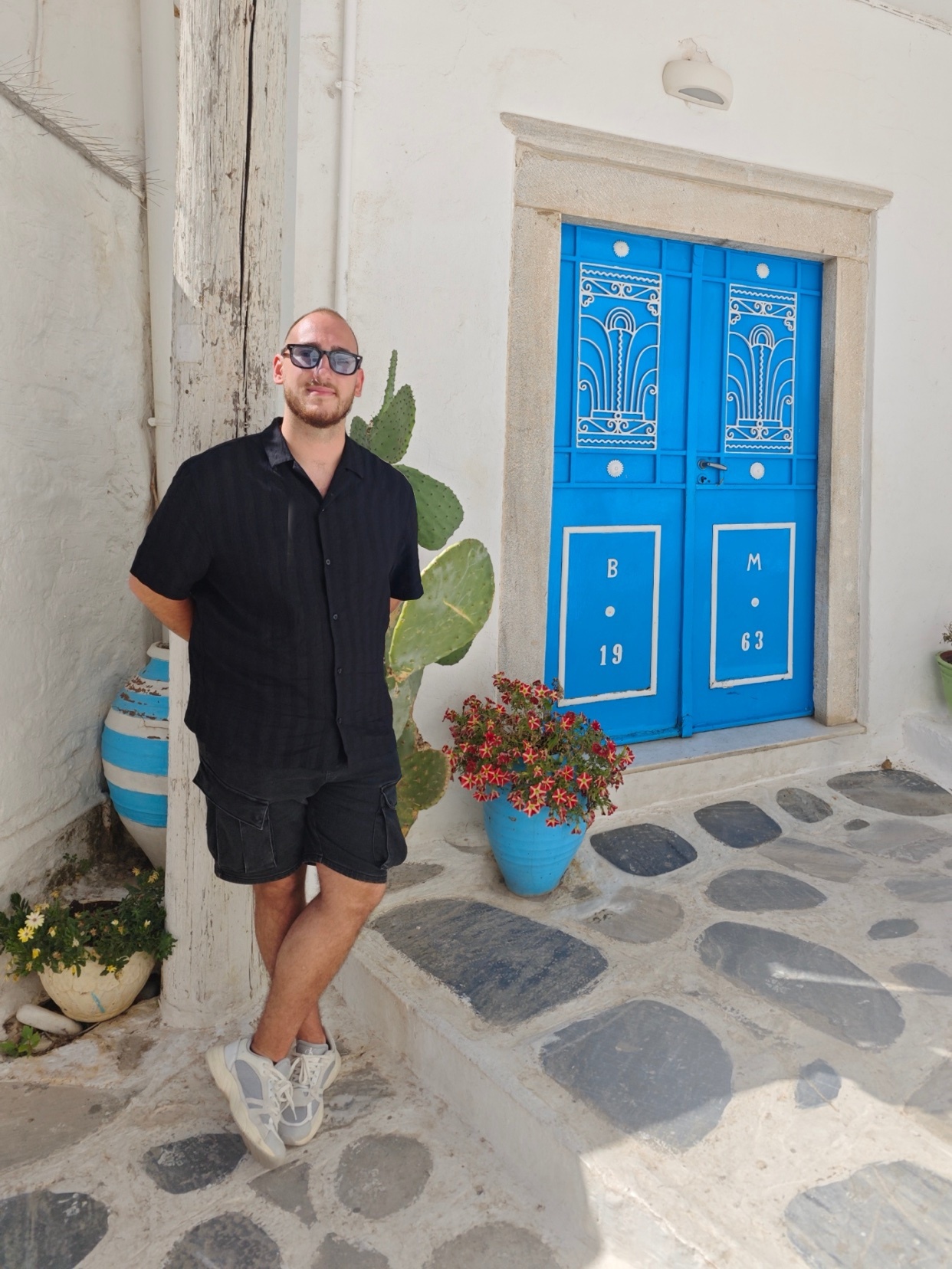 A man poses in front of a blue door. He is wearing sunglasses and a black shirt.