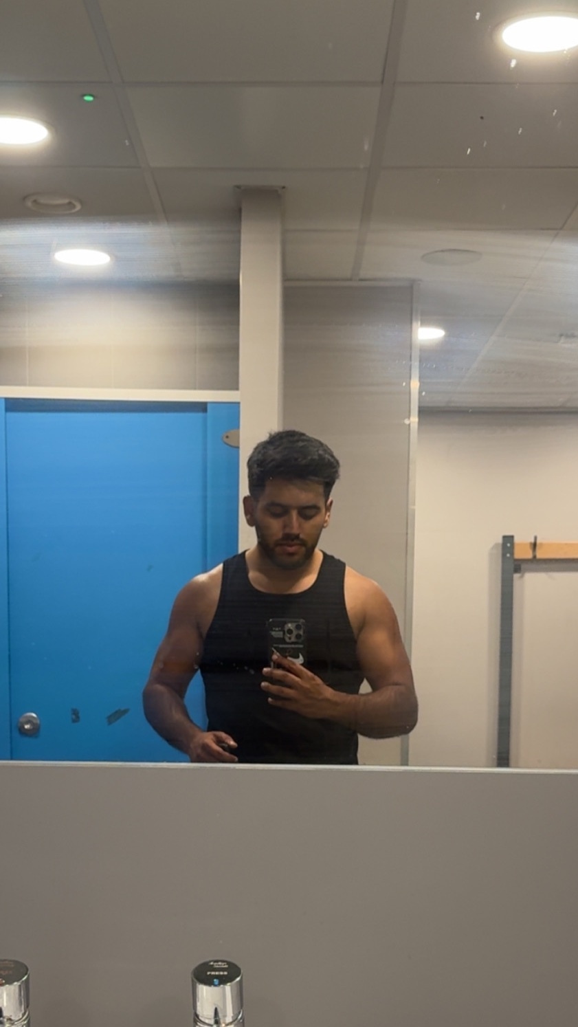 A man takes a mirror selfie in a gym locker room, wearing a black tank top and showing off his arms and chest. The reflection shows a blue door and gym equipment in the background.