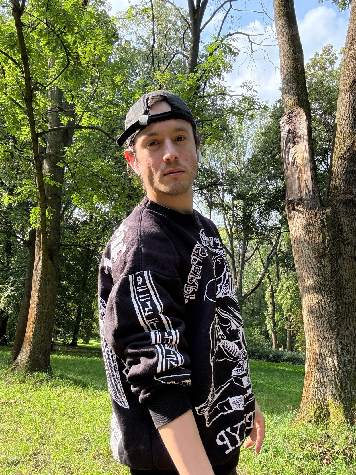 A man wearing a black graphic print crewneck sweatshirt and a black baseball cap poses for a photo in a park setting. He is looking towards the camera with a slight smile.