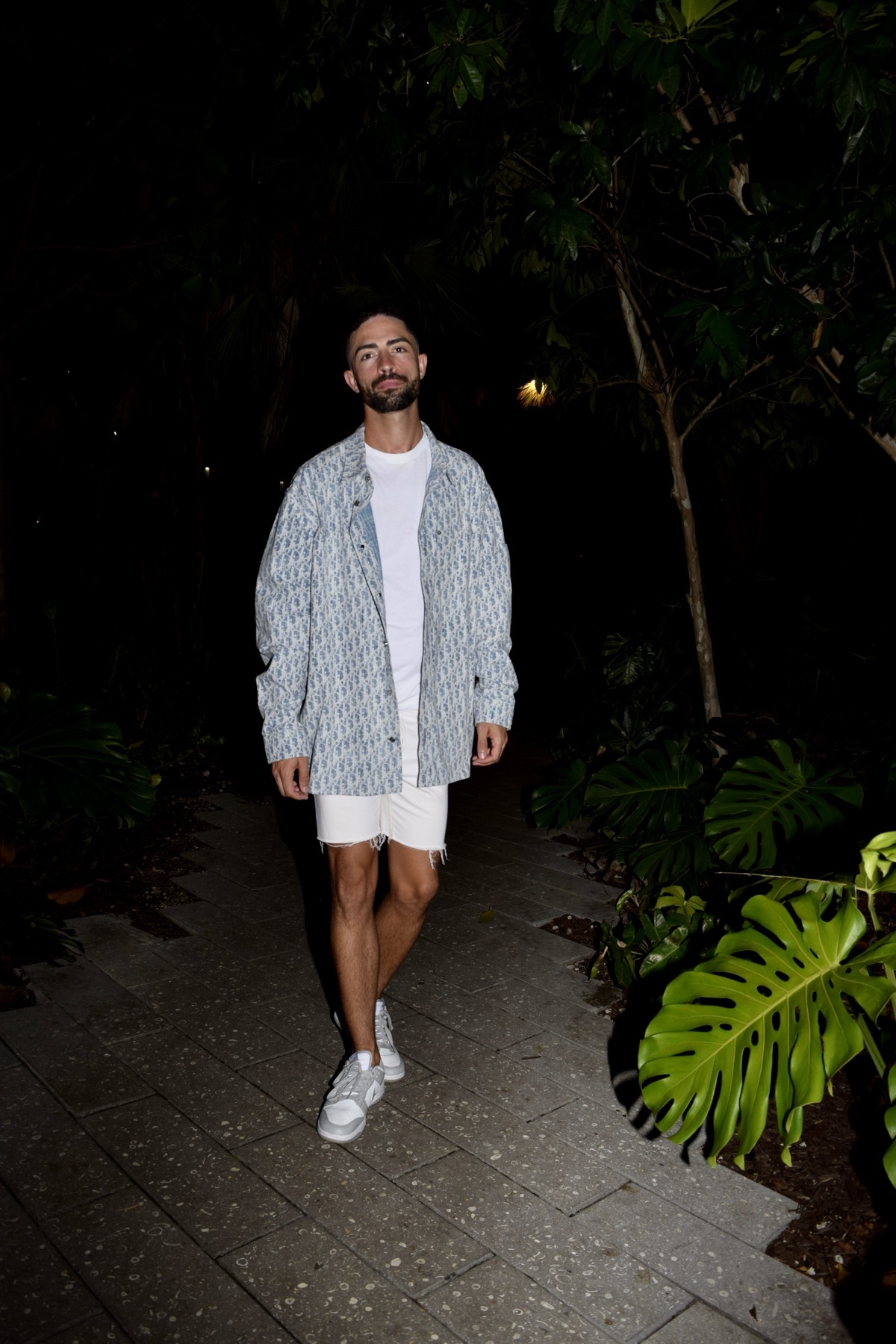 A man with a beard and short dark hair stands on a pathway at night, wearing a white t-shirt, white shorts, and an open button-up shirt. He is surrounded by lush green plants and trees.