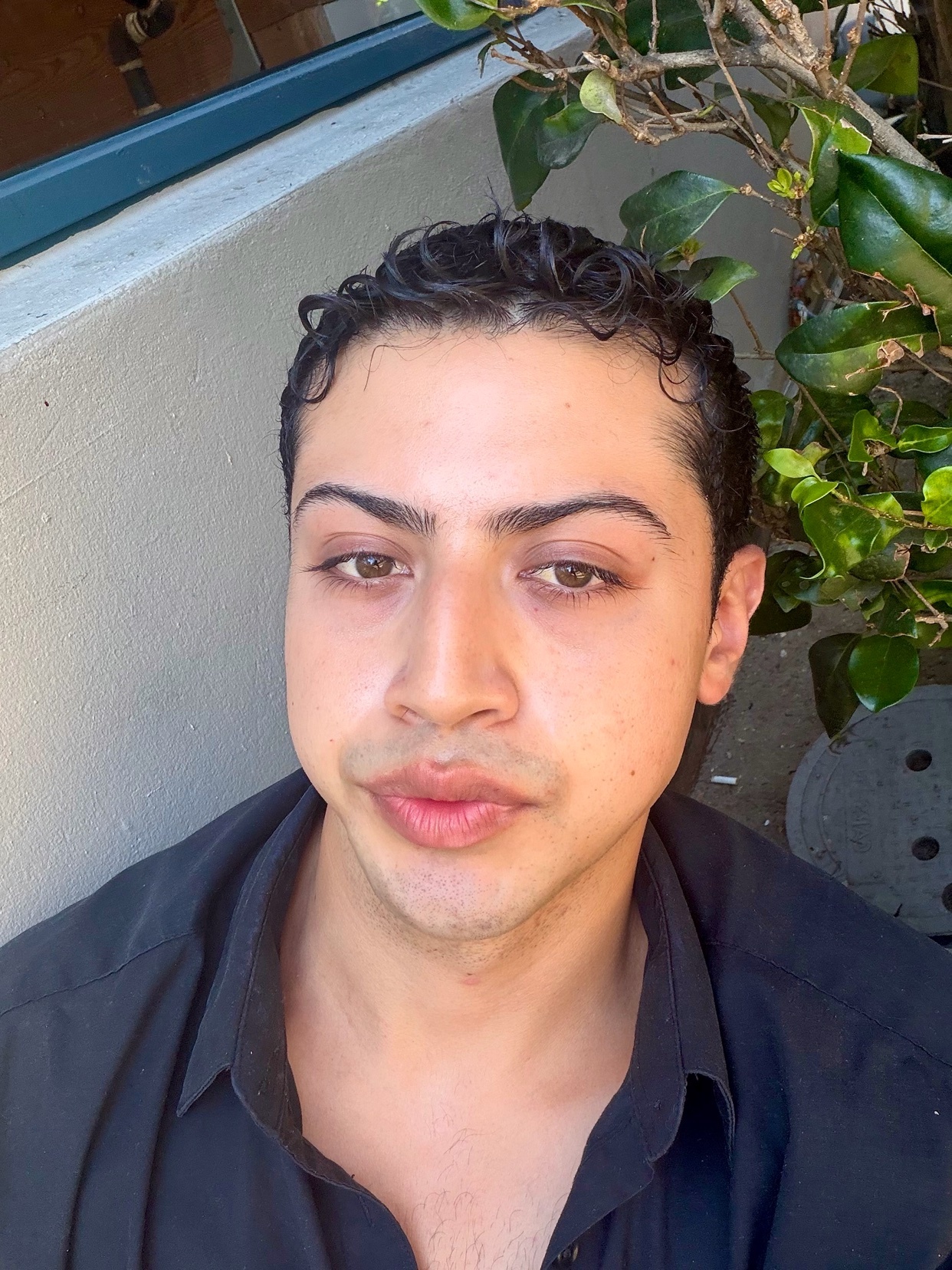 A close-up portrait of a man with dark, curly hair and defined eyebrows. He is wearing a black collared shirt and looking directly at the camera.