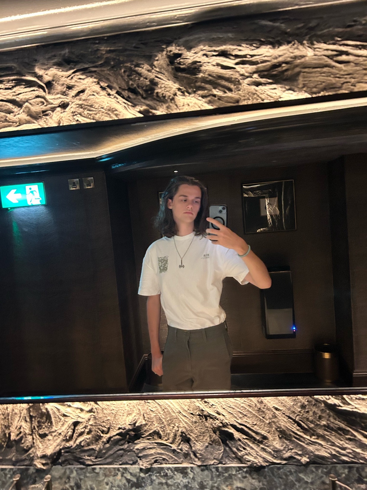 A young man with long, wavy brown hair takes a mirror selfie in a restroom. He is wearing a white t-shirt with a graphic design and olive green pants.