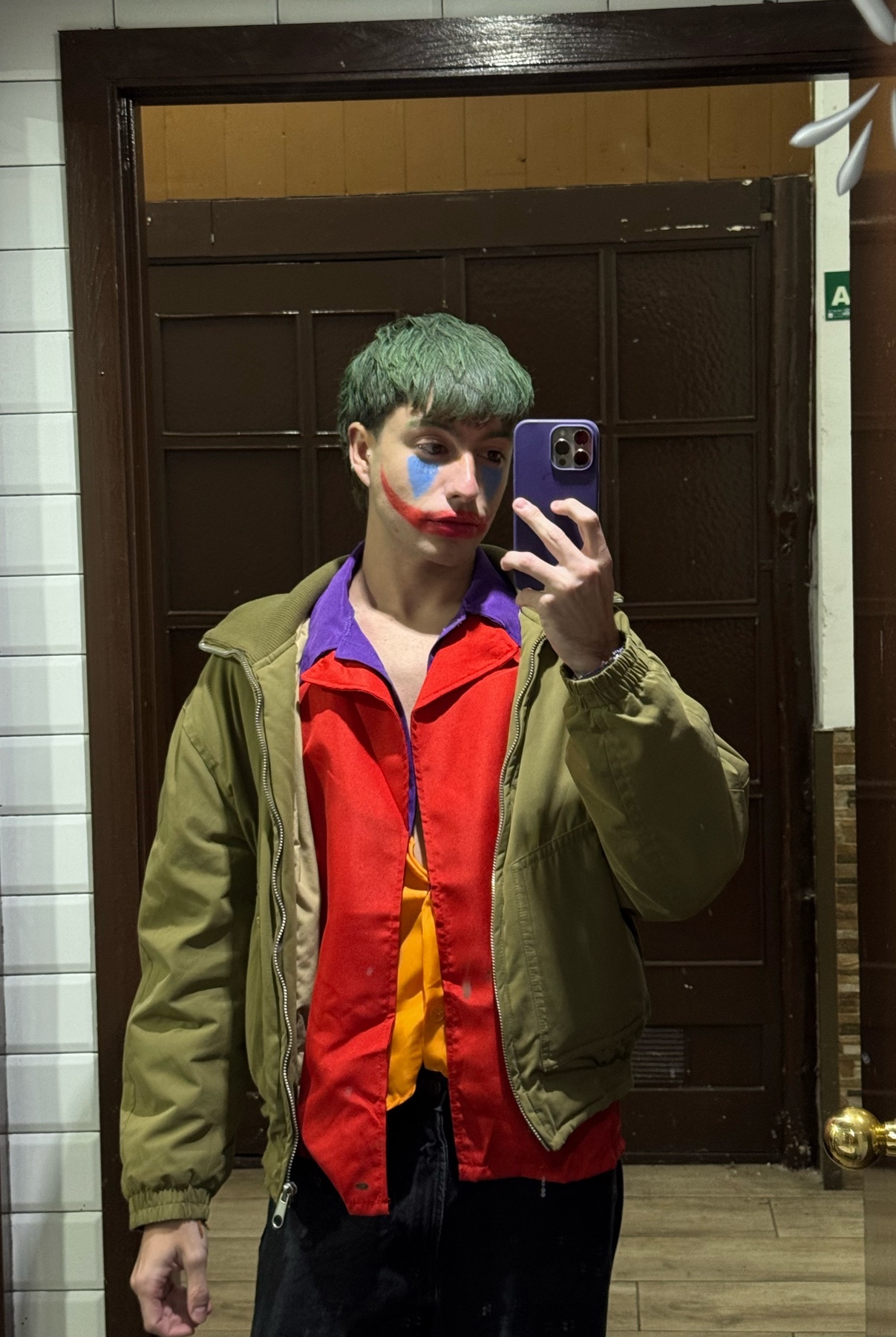 A man dressed as the Joker takes a mirror selfie. He is wearing makeup and a costume.