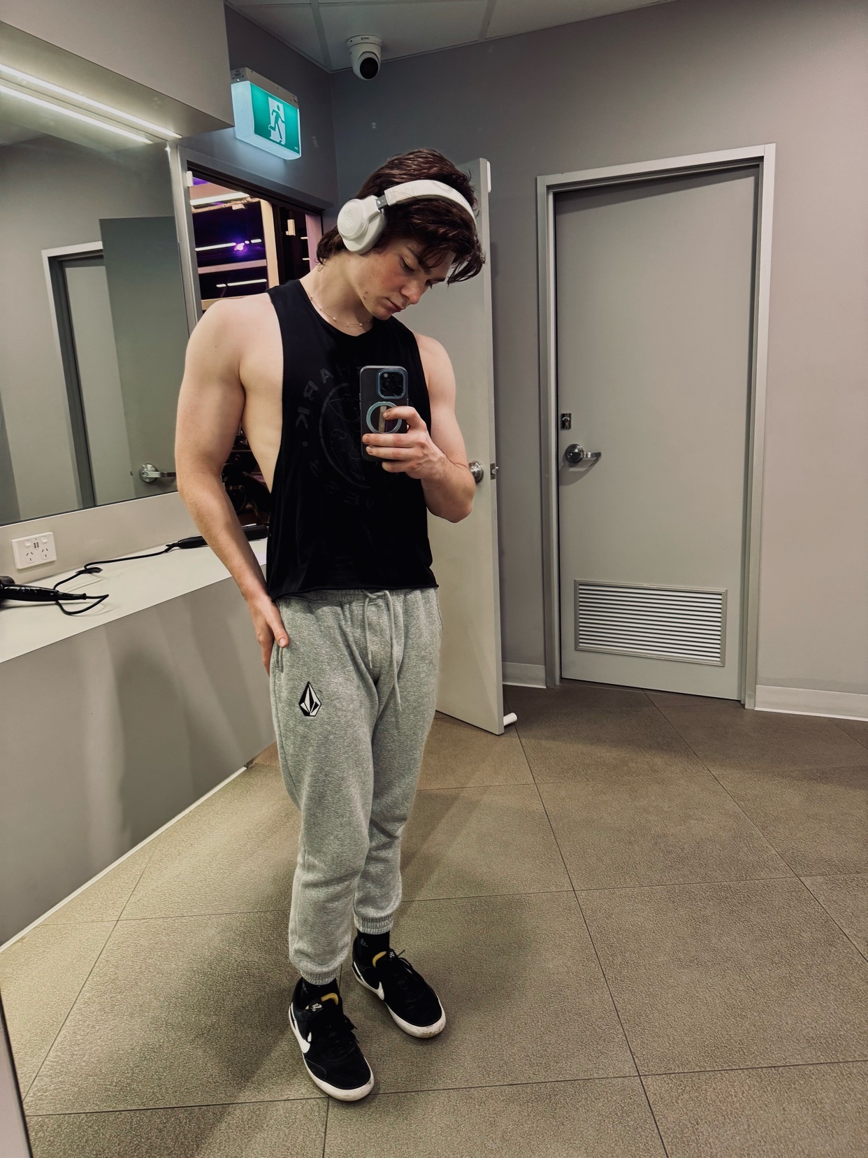 A young man in a black tank top and grey sweatpants takes a mirror selfie in a gym. He is wearing white headphones and black sneakers.