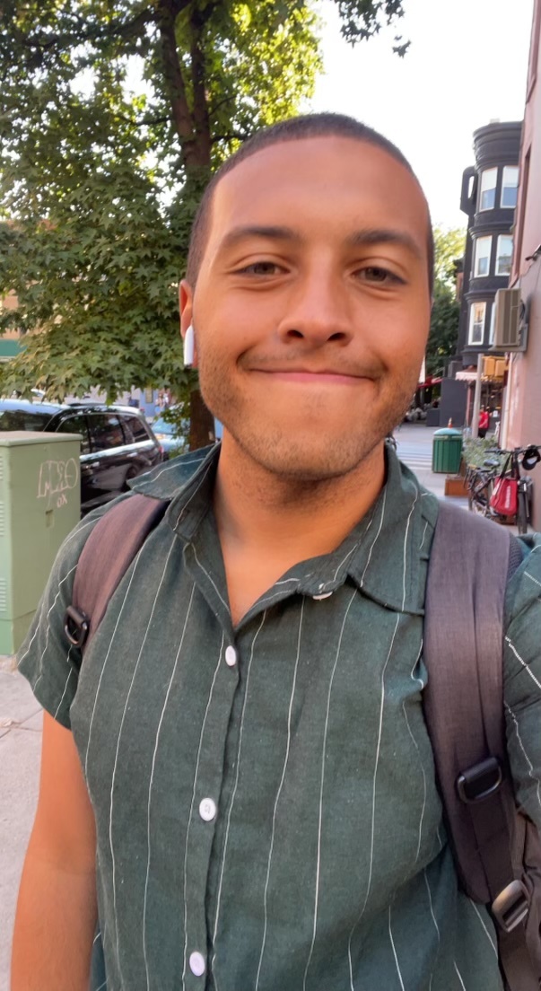 A man with a fresh haircut smiles at the camera while wearing earbuds and a green striped shirt. He has a backpack on and is standing outdoors with trees and buildings in the background.
