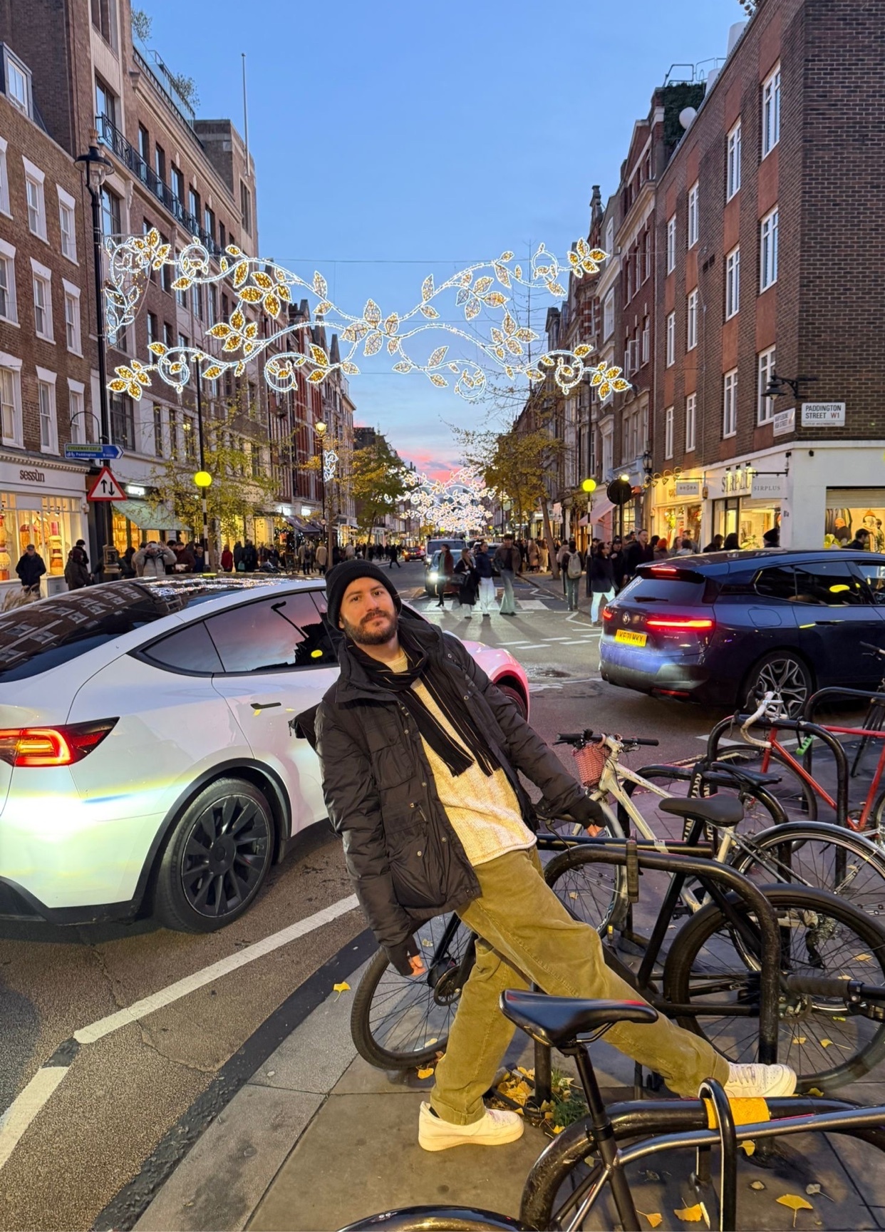A man poses on a bicycle rack on a city street. Christmas decorations hang overhead, and a Tesla is parked nearby.