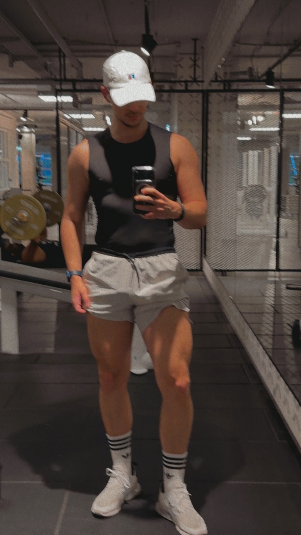 A man in athletic wear takes a mirror selfie in a gym. He is wearing a black tank top, grey shorts, a white baseball cap, and white sneakers with socks.