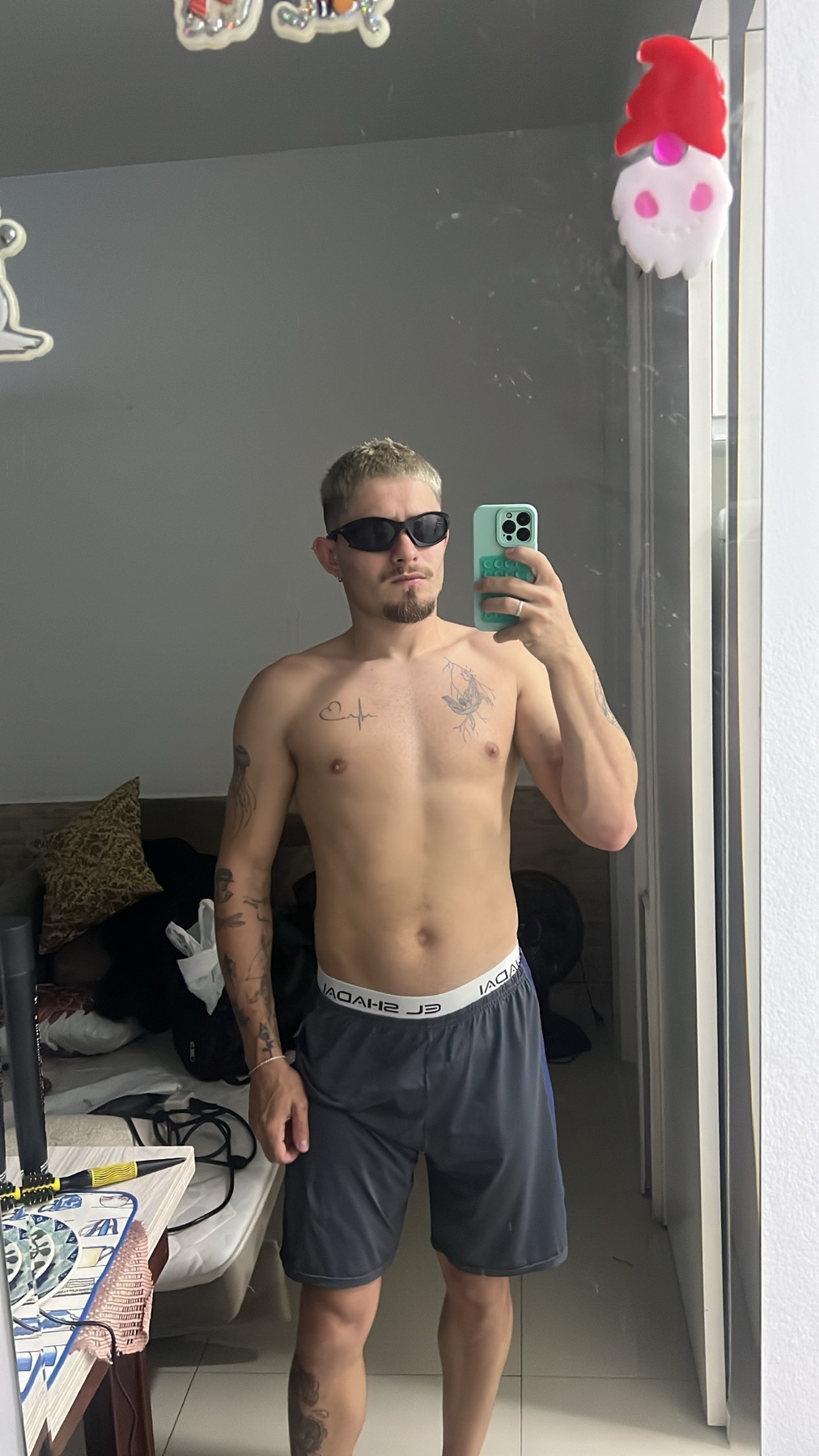 A man with blond hair and stubble takes a mirror selfie while wearing sunglasses and shorts. He is shirtless, showing off his tattoos.