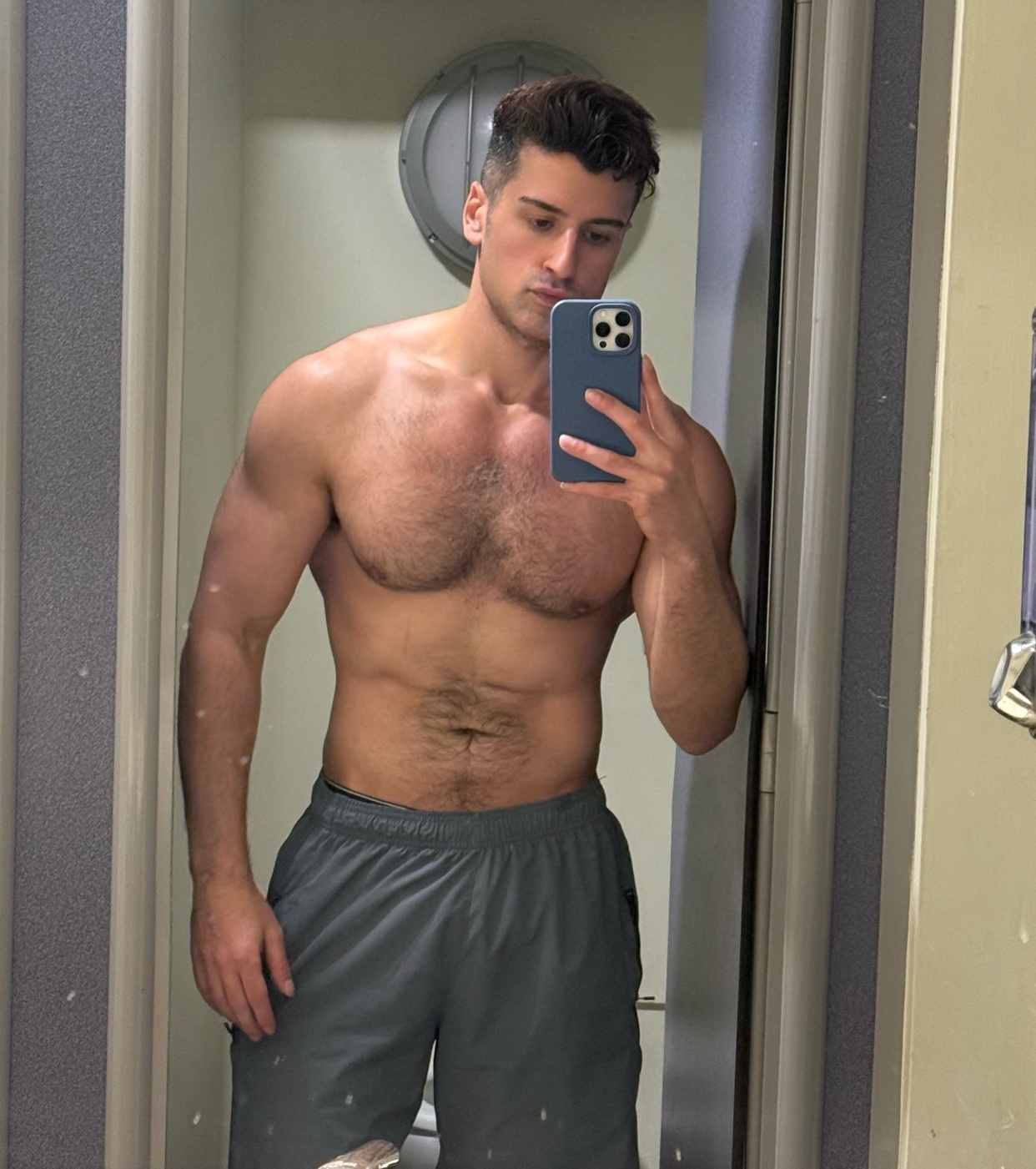 A man with dark hair and a muscular physique takes a selfie in a gym locker room. He is wearing grey athletic shorts and is shirtless, showing his chest and abs.