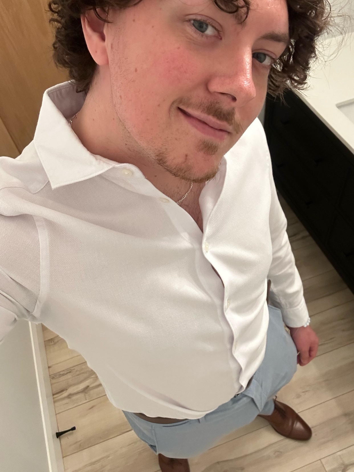 A man with curly hair and stubble poses for a selfie wearing a white button-down shirt, light blue dress pants, and brown dress shoes.