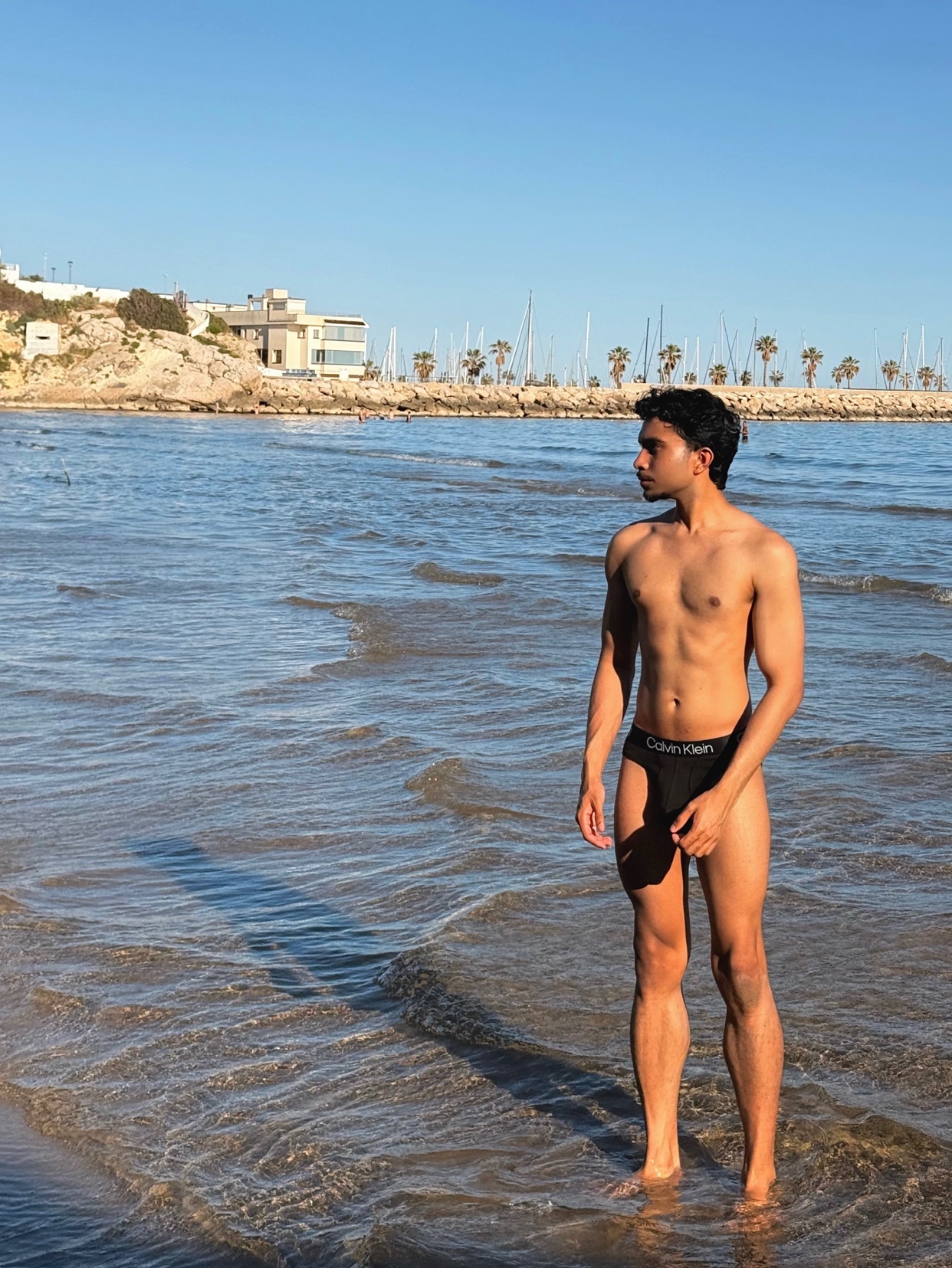 A shirtless man stands in the shallow ocean water at the beach, wearing black Calvin Klein underwear. He is looking out towards the coastline under a clear blue sky.
