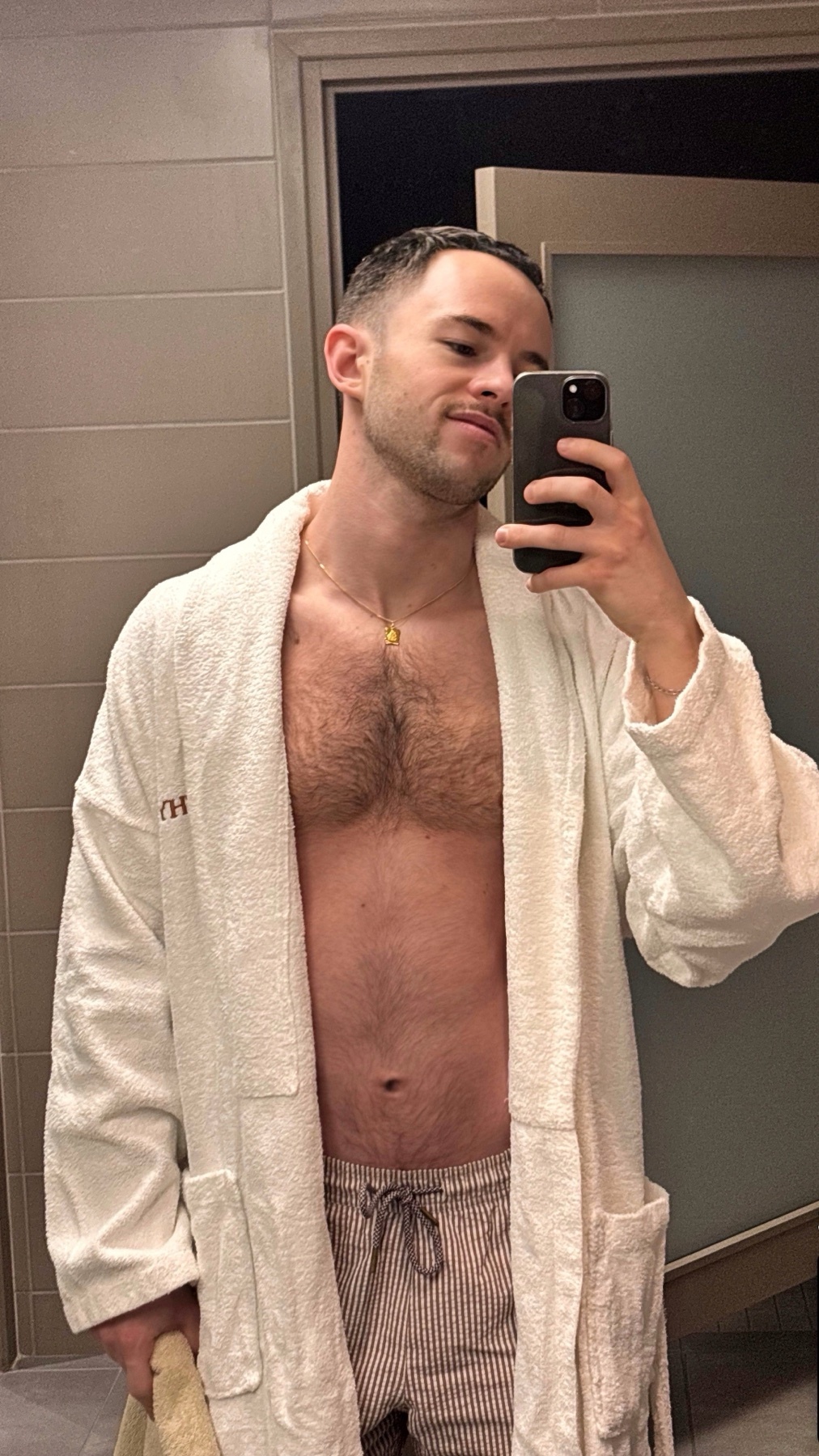 A man takes a selfie in a white bathrobe and pajama pants, showing off his chest hair and stubble.