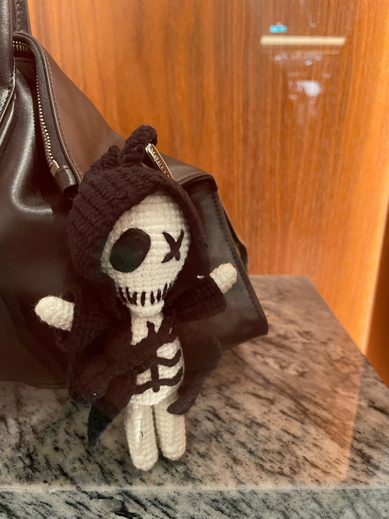 A close-up shot of a handmade crochet skeleton doll with a black hoodie. The doll is attached to a brown leather bag.