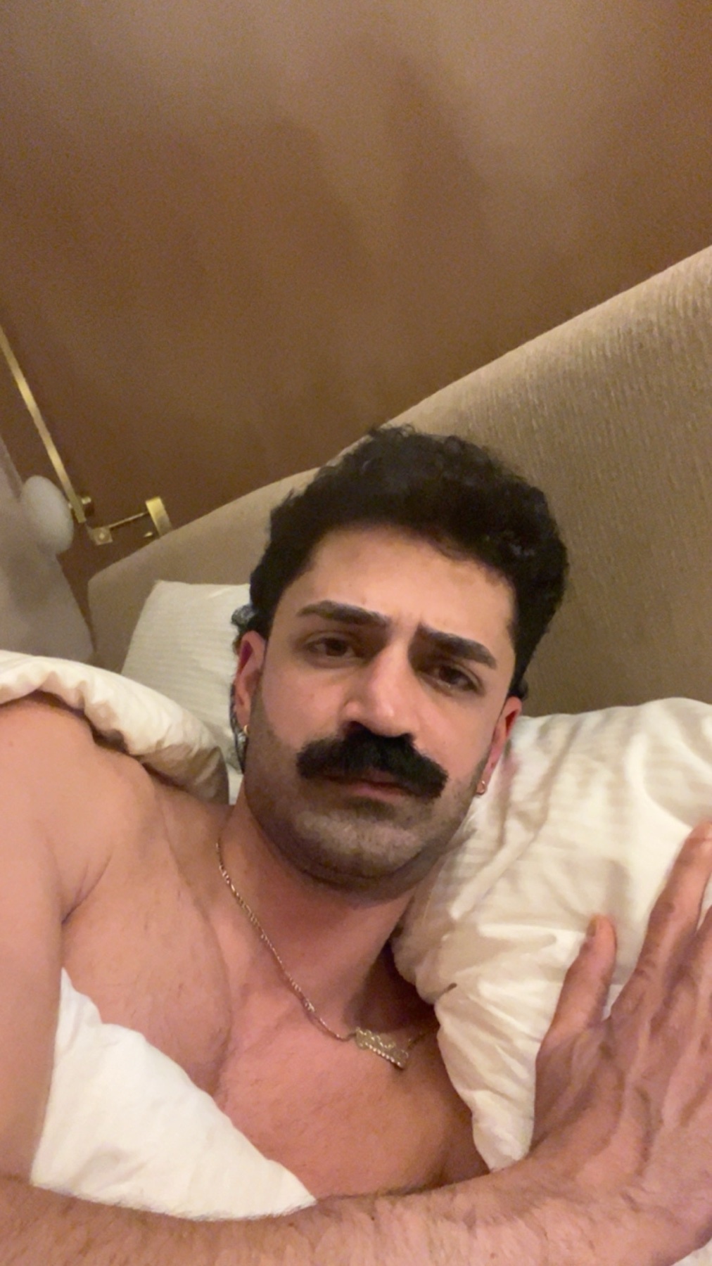 A man with a prominent mustache lies in bed, looking directly at the camera. He is partially covered by blankets and pillows.
