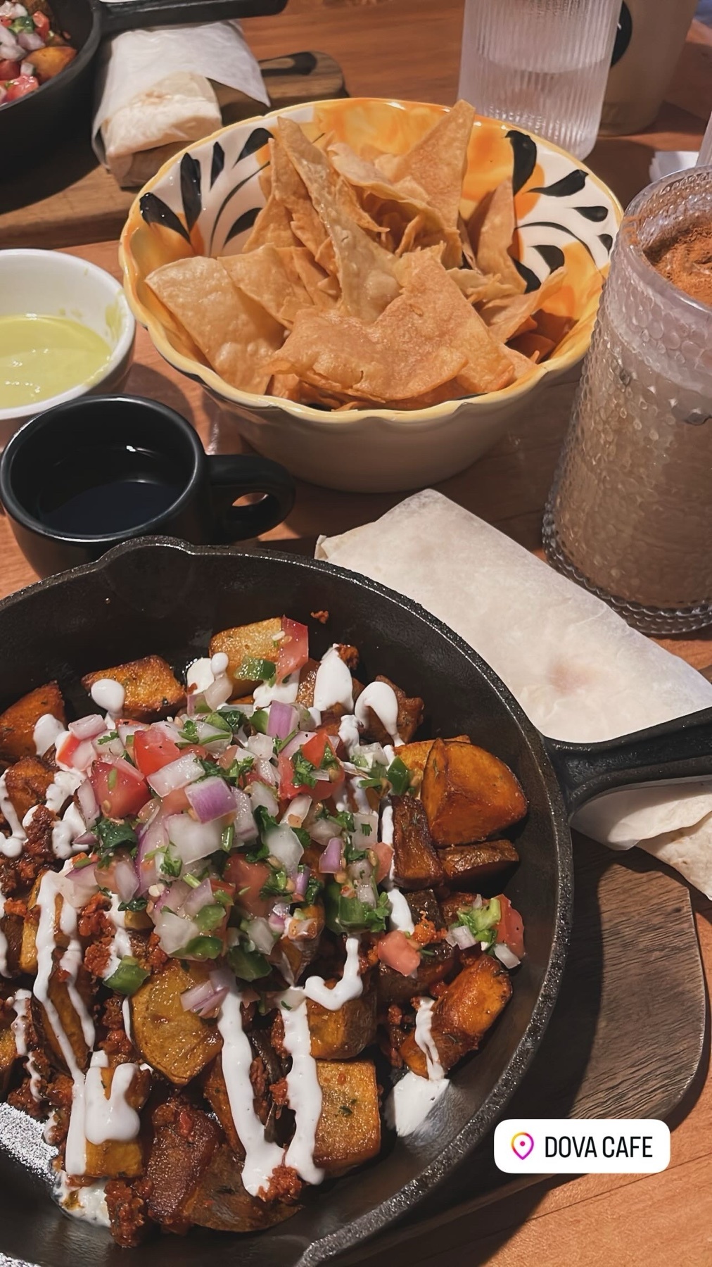 A close-up shot of a hearty breakfast dish served in a cast-iron skillet. The meal is topped with pico de gallo and a drizzle of white sauce, accompanied by a bowl of tortilla chips and dips.