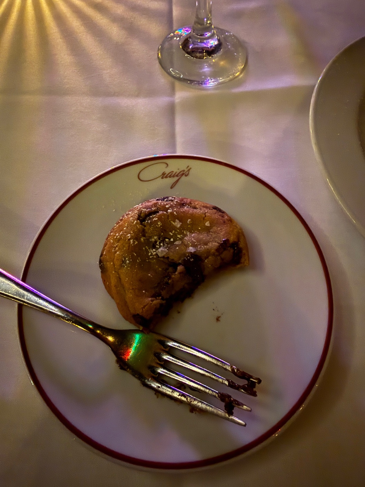 A close-up shot of a half-eaten chocolate chip cookie on a white plate with a maroon rim. A fork with crumbs rests on the plate, and a wine glass is visible in the background.