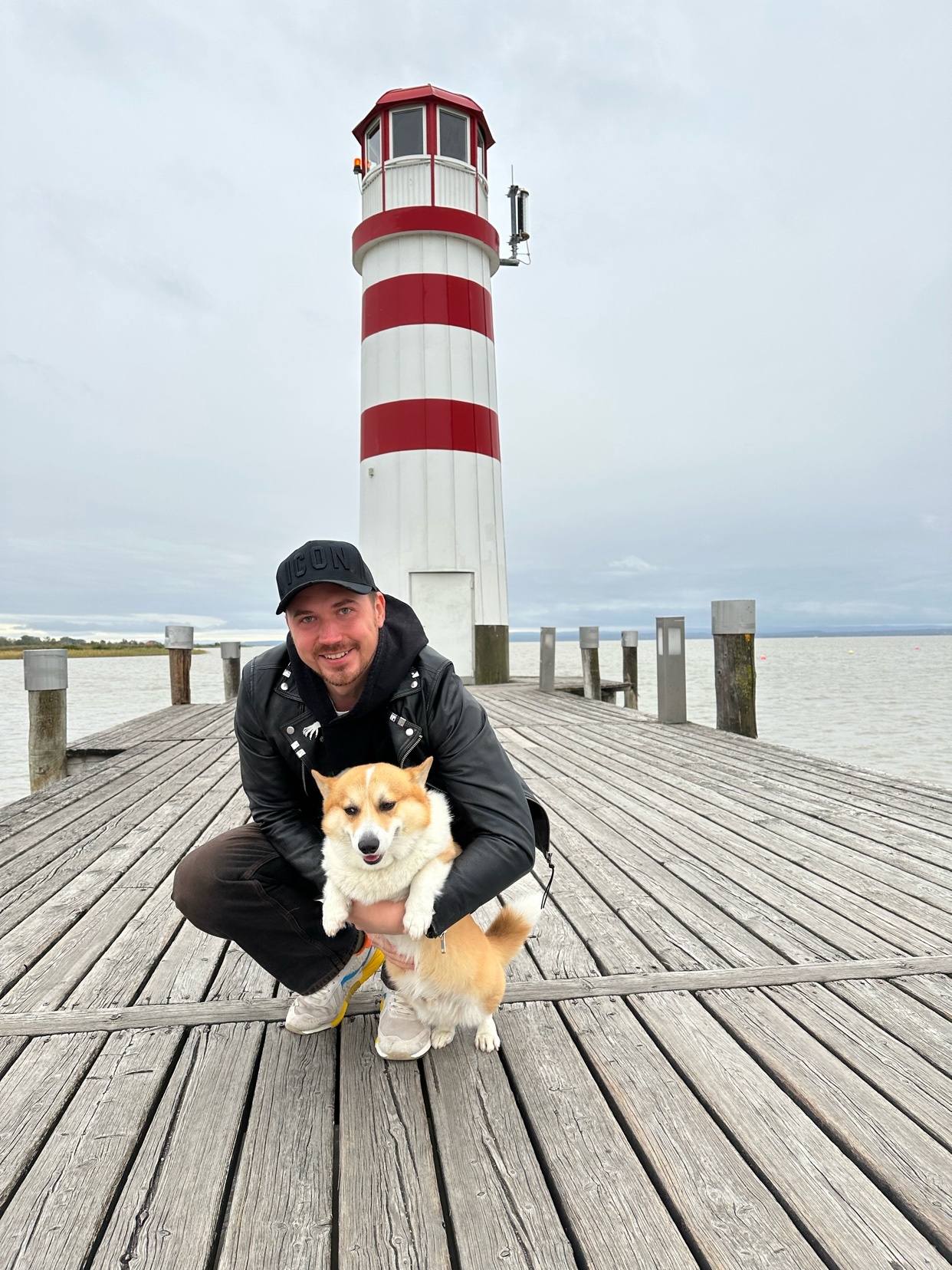 A man kneels on a wooden pier, holding a corgi dog. A lighthouse stands tall in the background.
