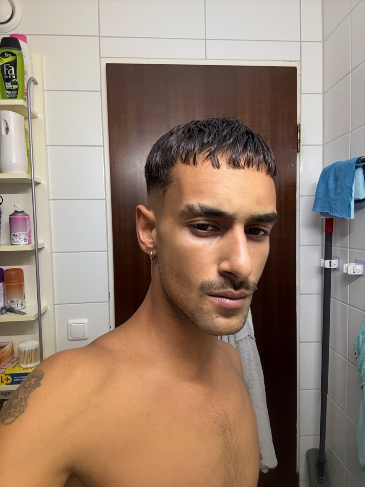 A man takes a shirtless selfie in a bathroom. He has dark hair and a mustache.