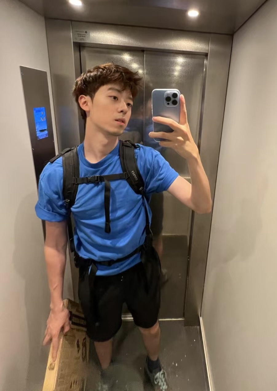 A young man takes a selfie in an elevator while wearing a blue t-shirt and a backpack.