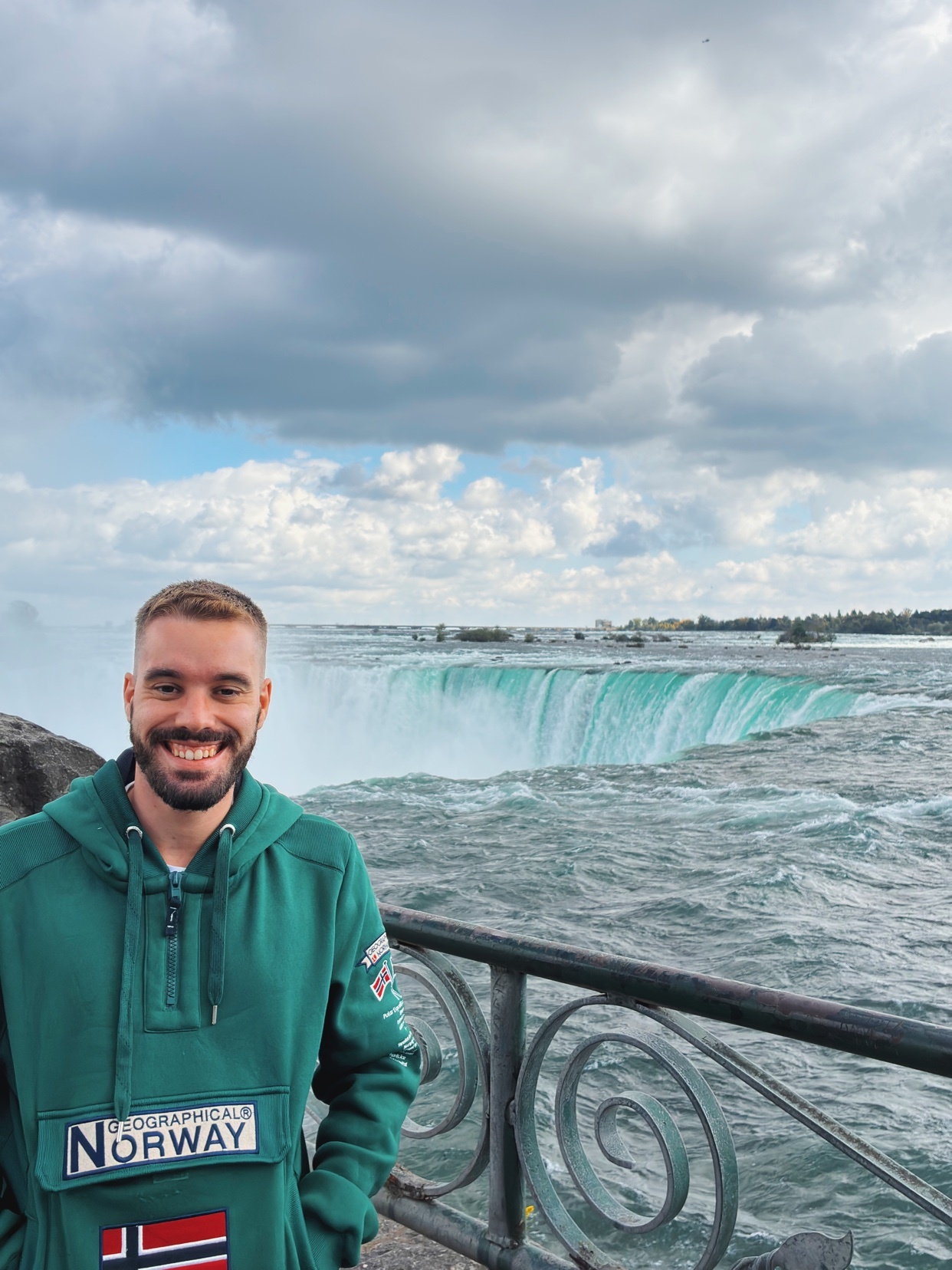 A man smiles in front of Niagara Falls. He is wearing a green hoodie and appears to be enjoying his trip.