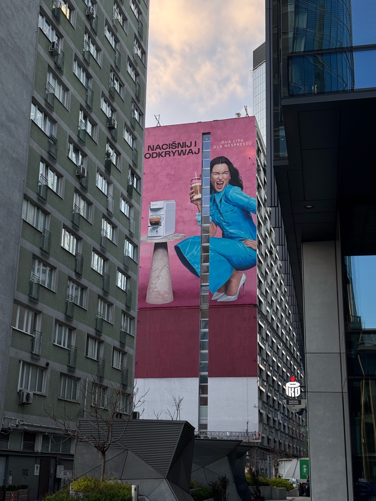 A large billboard features singer Dua Lipa in a vibrant blue outfit, crouching next to a small appliance. The advertisement is displayed on the side of a tall building in an urban setting.