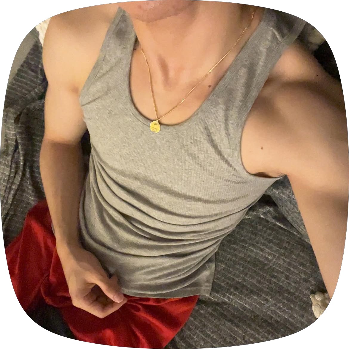 A man in a grey tank top and red shorts is lying down, showing off his muscular arms and chest. He is wearing a gold chain around his neck.
