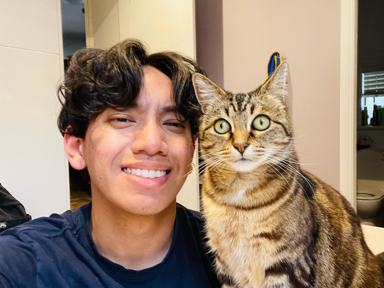 A man smiles for a selfie with his cat. The cat is a tabby, and the man has curly hair.