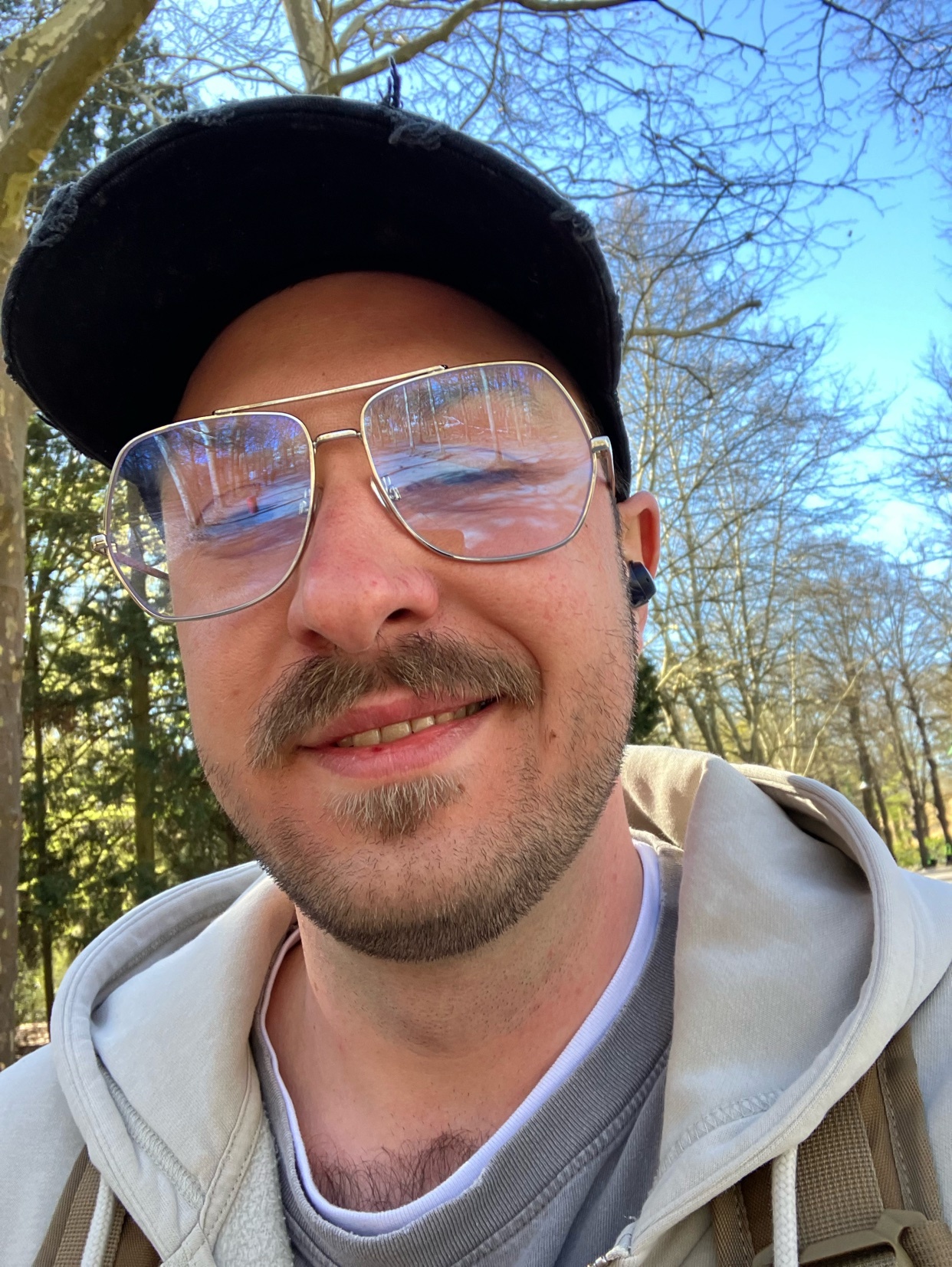A man wearing glasses and a baseball cap smiles for a selfie outdoors.