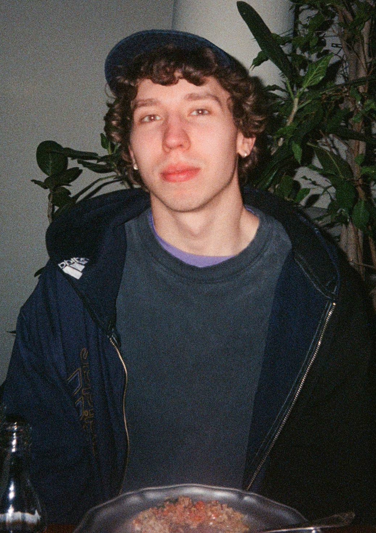 A young man with curly brown hair and a baseball cap poses for a photo indoors. He is wearing a dark t-shirt and a jacket.