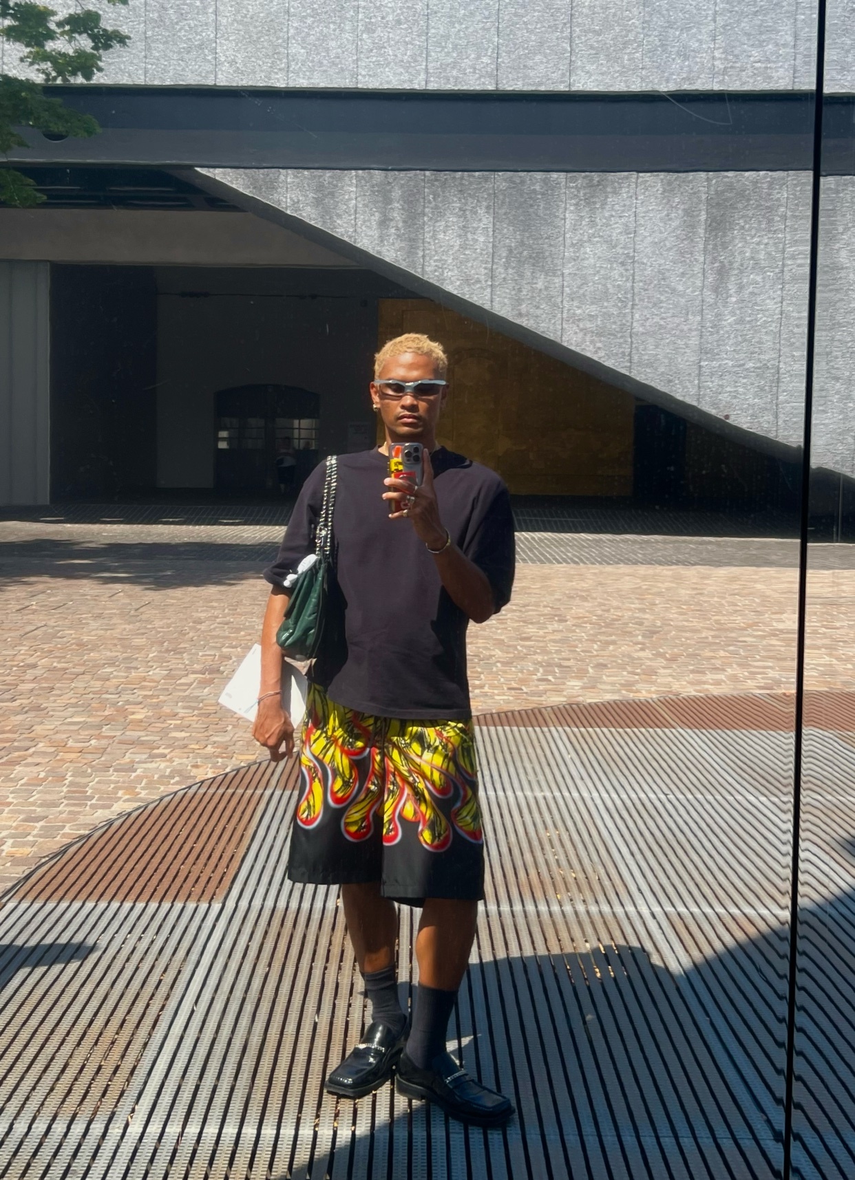 A man with blond hair and sunglasses poses for a mirror selfie in a black t-shirt and bold flame print shorts. He is wearing black socks and loafers, and carrying a green bag.