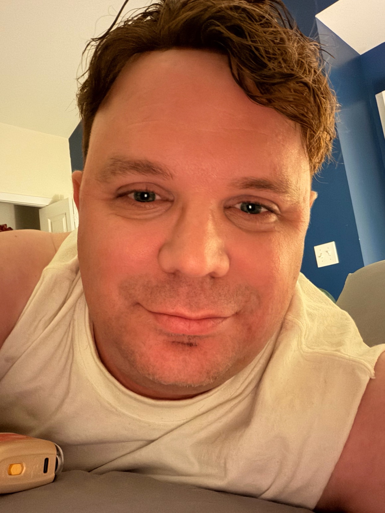 A man takes a selfie while getting ready for bed. He is wearing a white tank top and has a slight smile.